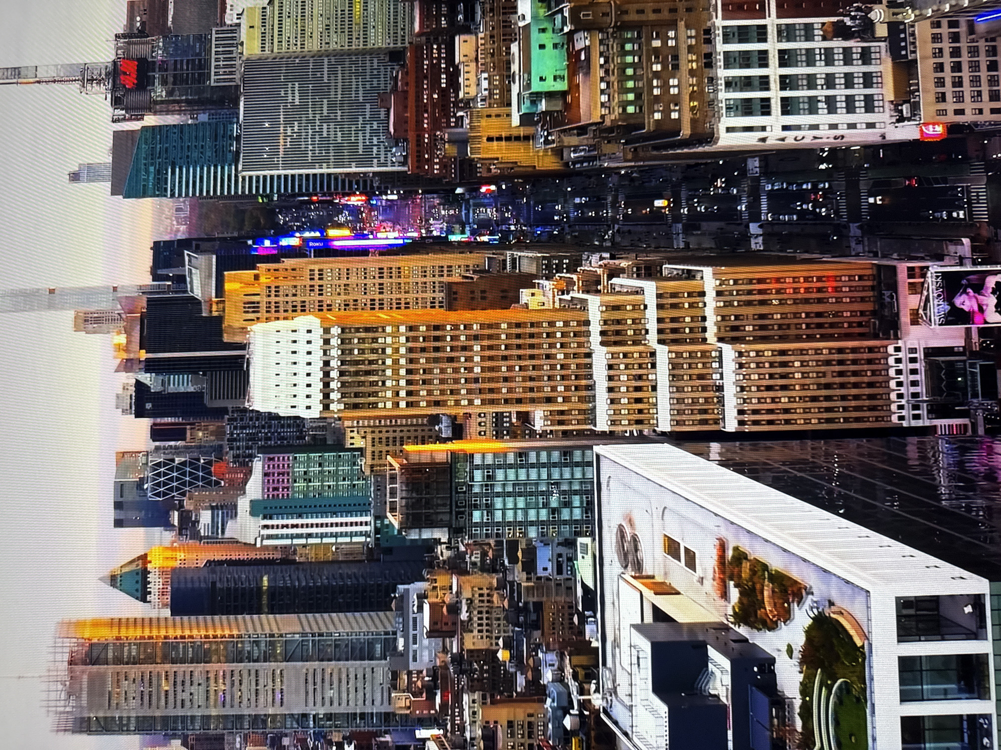 This image depicts a dense urban skyline with numerous skyscrapers. The buildings vary in architectural style and color, with some featuring glass facades and others made of brick or concrete. The scene is bathed in the warm light of either sunrise or sunset, casting long shadows and highlighting the textures of the buildings. A street lined with cars and illuminated by neon signs runs vertically through the center of the frame, suggesting a busy commercial district. Several buildings are adorned with illuminated signage, including one for "SWAROVSKI." The presence of a large green rooftop garden on a lower building adds a touch of nature amidst the concrete jungle. The overall impression is that of a bustling metropolis.
This image is not of Burgas, Bulgaria. The architecture and urban landscape are characteristic of New York City, likely Midtown Manhattan, given the density of skyscrapers, the illuminated street scene resembling Times Square, and the presence of iconic building styles.