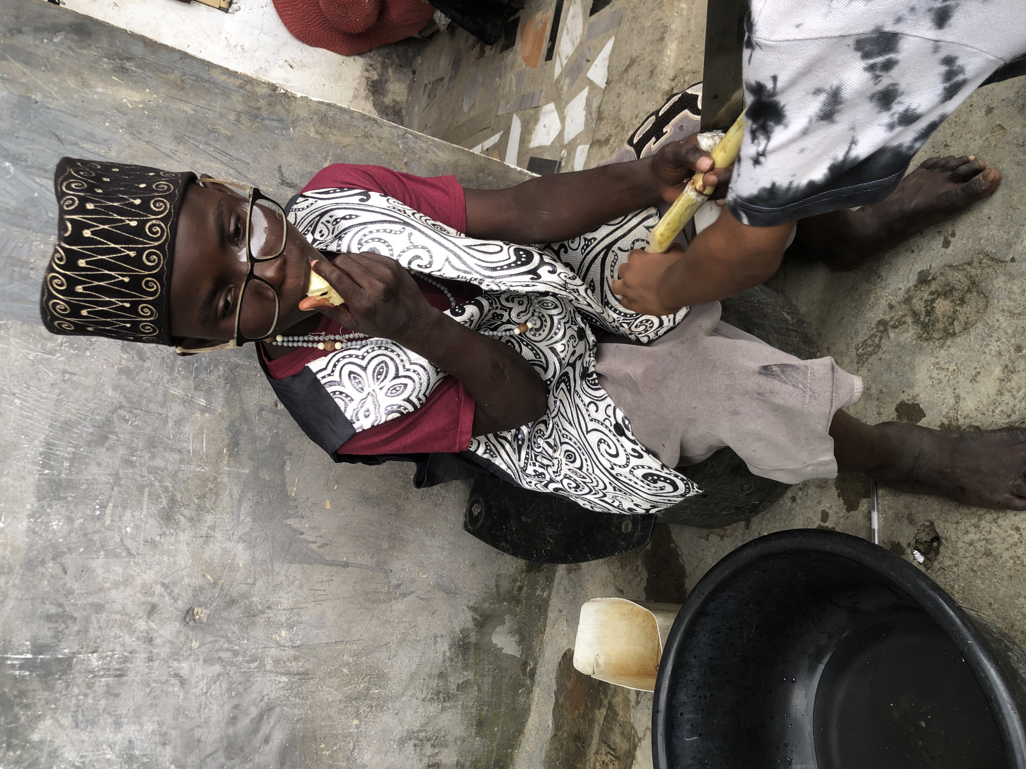 In Ado, Nigeria, a young boy, wearing a black and gold patterned Kufi, glasses, and a maroon shirt with a black and white patterned vest, sits on a black tire. He is eating a piece of fruit. Another person, whose hands are visible, is peeling a piece of sugarcane for him. A large black basin filled with water is to the boy's left, and a small white plastic container is next to it. The scene takes place outdoors against a rough, grey concrete wall. The ground is concrete with some scattered debris.