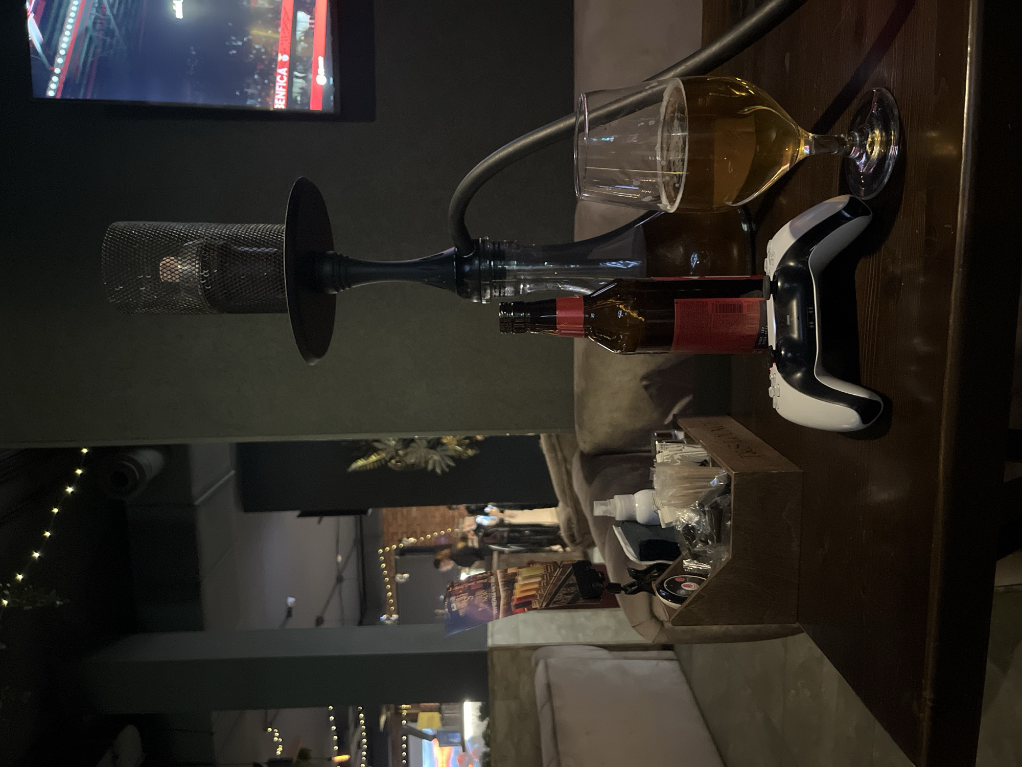 An overhead shot, viewed from an eye-level perspective looking down onto a wooden table, captures a dimly lit indoor setting, likely a bar or lounge in the evening.
In the foreground, a prominent black hookah with a mesh top and dark stem stands on the table. A flexible black hose extends from the hookah. Beside it are several brown glass bottles, one of which appears to have a red label. A clear wine glass containing an amber-colored liquid, possibly beer, is tilted on its side near the bottles. To the right of the glass, a white and black gaming controller, resembling a PlayStation DualSense, rests on the table. A small, dark wooden caddy filled with various small containers, possibly hand sanitizers or other amenities, is also visible on the table to the right.
In the background, a large television screen displays what appears to be a sports match, with the word "BENFICA" clearly visible, suggesting a soccer game involving the Portuguese club S.L. Benfica. Below the TV and extending further into the background, other parts of the lounge are visible, characterized by soft, warm string lights, blurred figures of people, and comfortable seating, creating a relaxed and social atmosphere. The overall scene suggests a casual entertainment space where patrons can enjoy drinks, hookah, gaming, and sports.