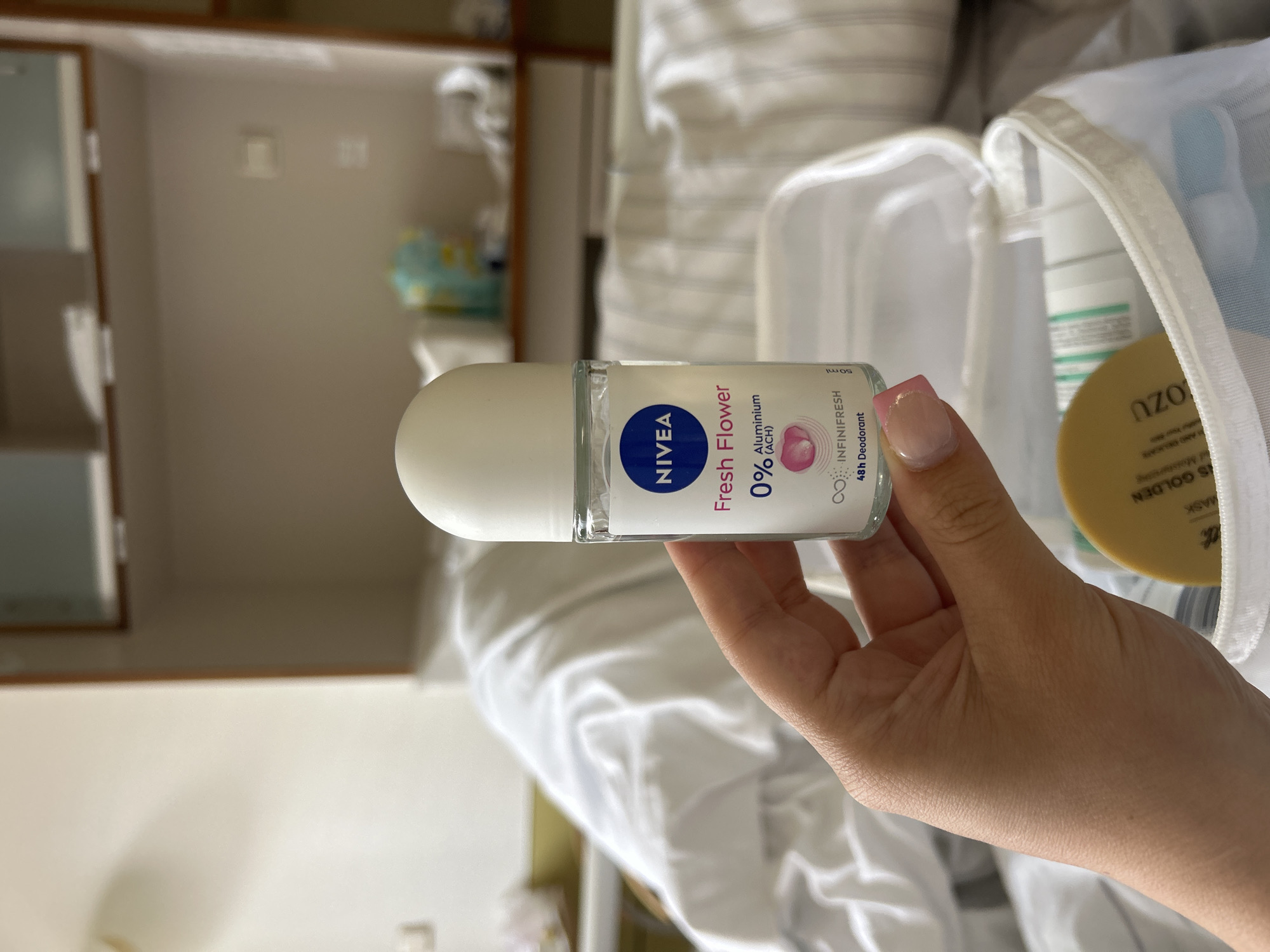 A person's hand holds a Nivea Fresh Flower 0% Aluminium (ACH) roll-on deodorant. The product is in a clear bottle with a white cap, and the label indicates it provides 48h Deodorant protection. The background suggests an indoor setting, possibly a hospital room, with white bedding and shelves visible in the distance. The person's fingernails are painted pink with a subtle glitter accent.