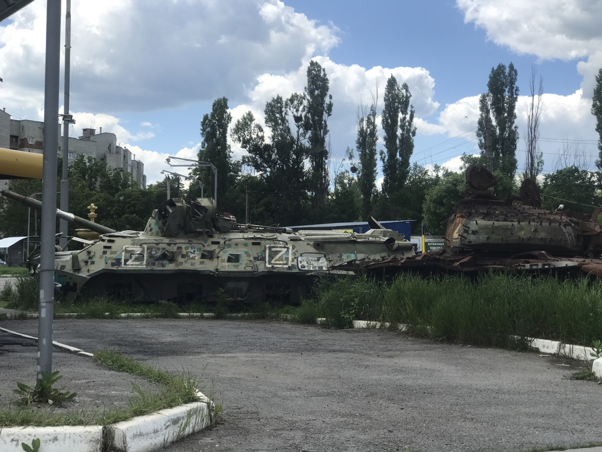 The image displays an outdoor scene featuring two heavily damaged or destroyed military vehicles under a partly cloudy sky. In the foreground on the left, a tall, grey utility pole stands. The ground consists of grey asphalt with patches of green grass growing through cracks and along a white curb line.
The primary subject on the left is a camouflaged armored personnel carrier (APC) or infantry fighting vehicle (IFV). Its paint scheme includes shades of green, brown, and possibly light blue/grey. A prominent white "Z" symbol is painted on its side, a common identifier for Russian forces. The vehicle appears damaged, with parts of its superstructure and armor dislodged or missing, and its main gun barrel points towards the left.
To the right of the APC, a more severely damaged and rusted vehicle, likely a tank or another armored vehicle, sits partially obscured by tall grass. Its turret appears to be heavily compromised or blown off, showing considerable structural damage and a rusty, burnt-out appearance.
In the background, a line of tall, slender trees, possibly poplars, runs across the scene. Behind these trees, distant grey residential buildings, possibly apartment blocks, are visible on the left. The sky is bright blue with scattered white, fluffy clouds, indicating daytime. A partially obscured sign in Ukrainian is visible behind the right vehicle, which reads "СЕРТИФІКАЦІЯ ТЕХ..." (CERTIFICATION TECH...).
The overall scene suggests a collection of war-damaged military equipment, possibly an open-air exhibition or a temporary storage site, reflecting the aftermath of a conflict.