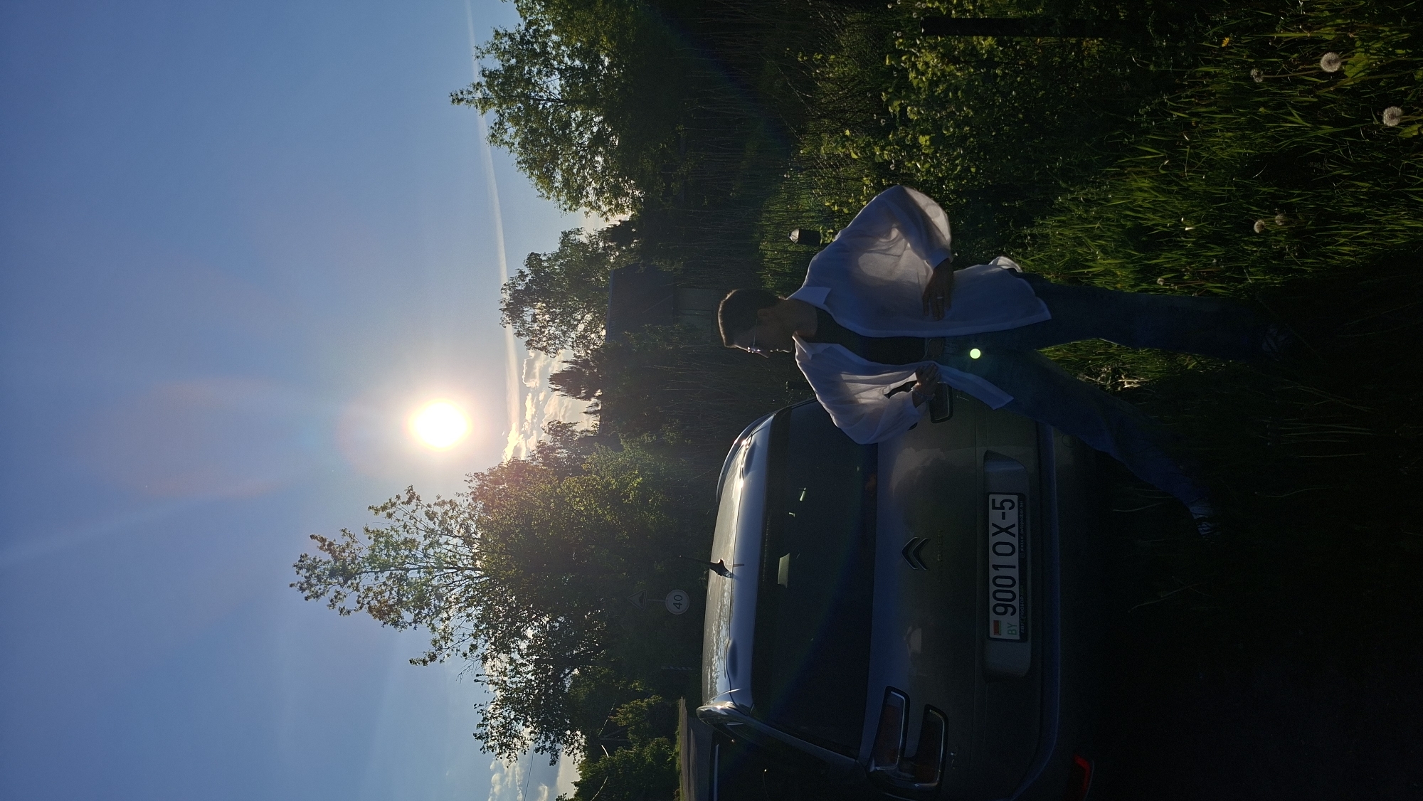 A person is leaning against the back of a silver Citroen car in a rural area near Сваткаўскі сельскі Савет, Belarus. The person is wearing a white shirt and jeans. The license plate of the car reads "9001OX-5". The scene is brightly lit by the sun. Trees and grass are visible in the background.