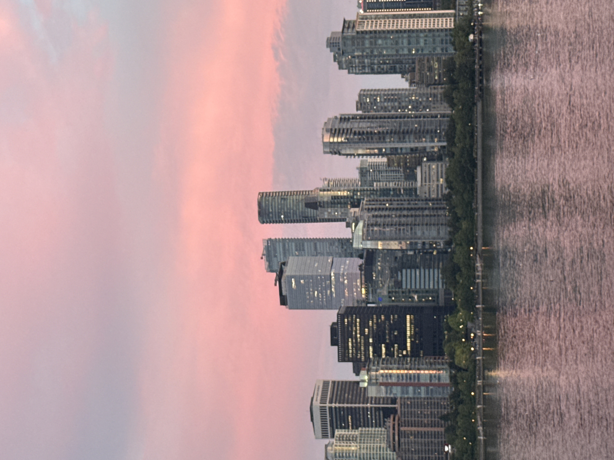 The image captures a panoramic view of the Vancouver skyline at dusk, with the sky painted in soft shades of pink and pale blue. The city's impressive array of modern skyscrapers dominates the scene, their windows reflecting the twilight hues. In the foreground, a calm body of water, likely the Burrard Inlet, mirrors the city lights and the sky's ethereal colors. A line of lush green trees separates the water from the urban landscape, with subtle lights visible along the waterfront promenade, suggesting the presence of people enjoying the evening. The overall impression is one of urban beauty juxtaposed with the tranquility of nature, characteristic of Vancouver's waterfront vistas.