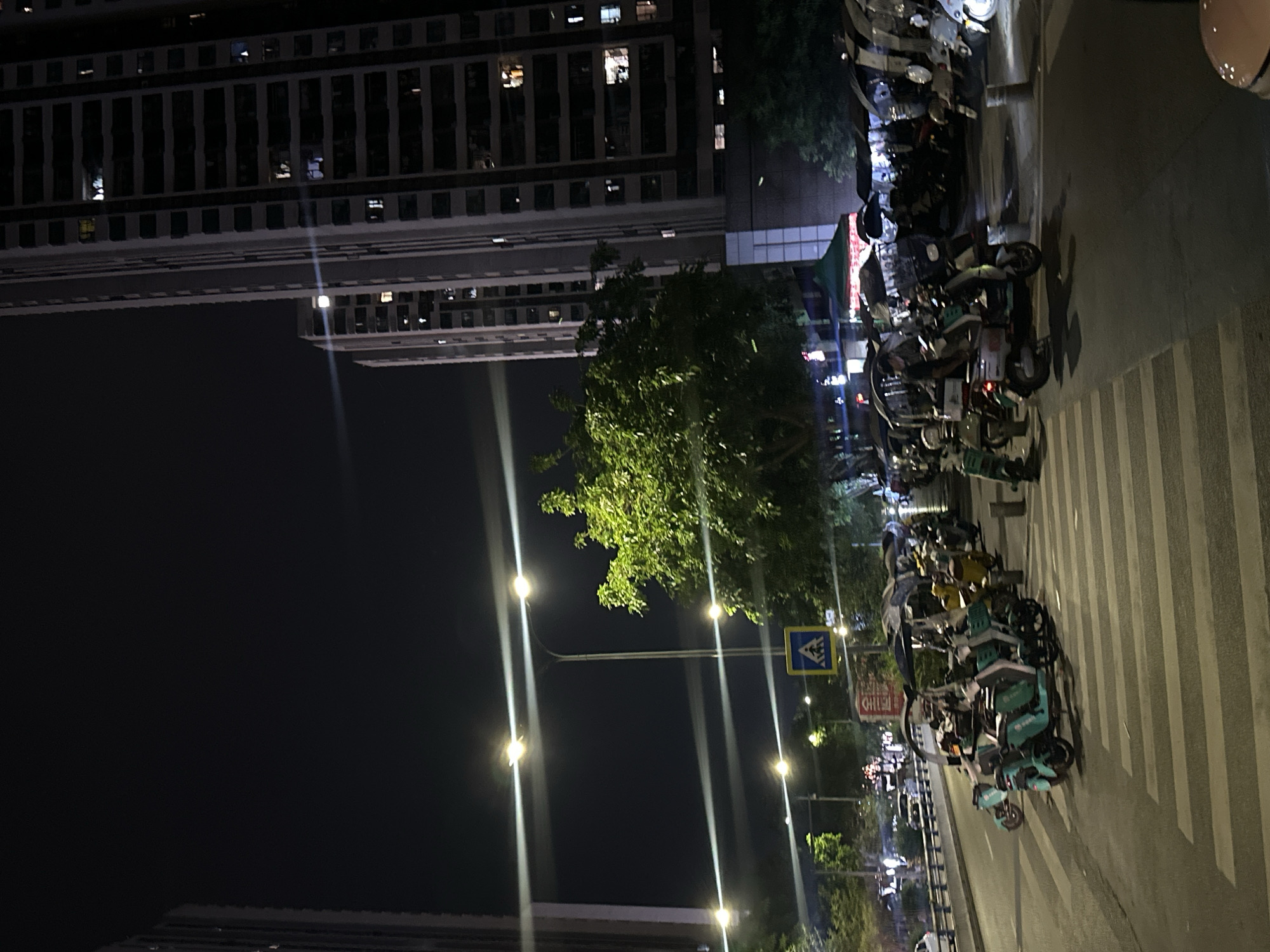 The image depicts a nighttime street scene in what appears to be an urban environment. A tall, multi-story building dominates the left side of the frame. Streetlights illuminate the scene, casting bright vertical beams of light. A dense green tree is visible between the building and the street. A row of parked electric scooters is lined up along the side of the road, next to a crosswalk. A street sign is also visible near the scooters. The location is likely Chongqing High-tech Zone, China.