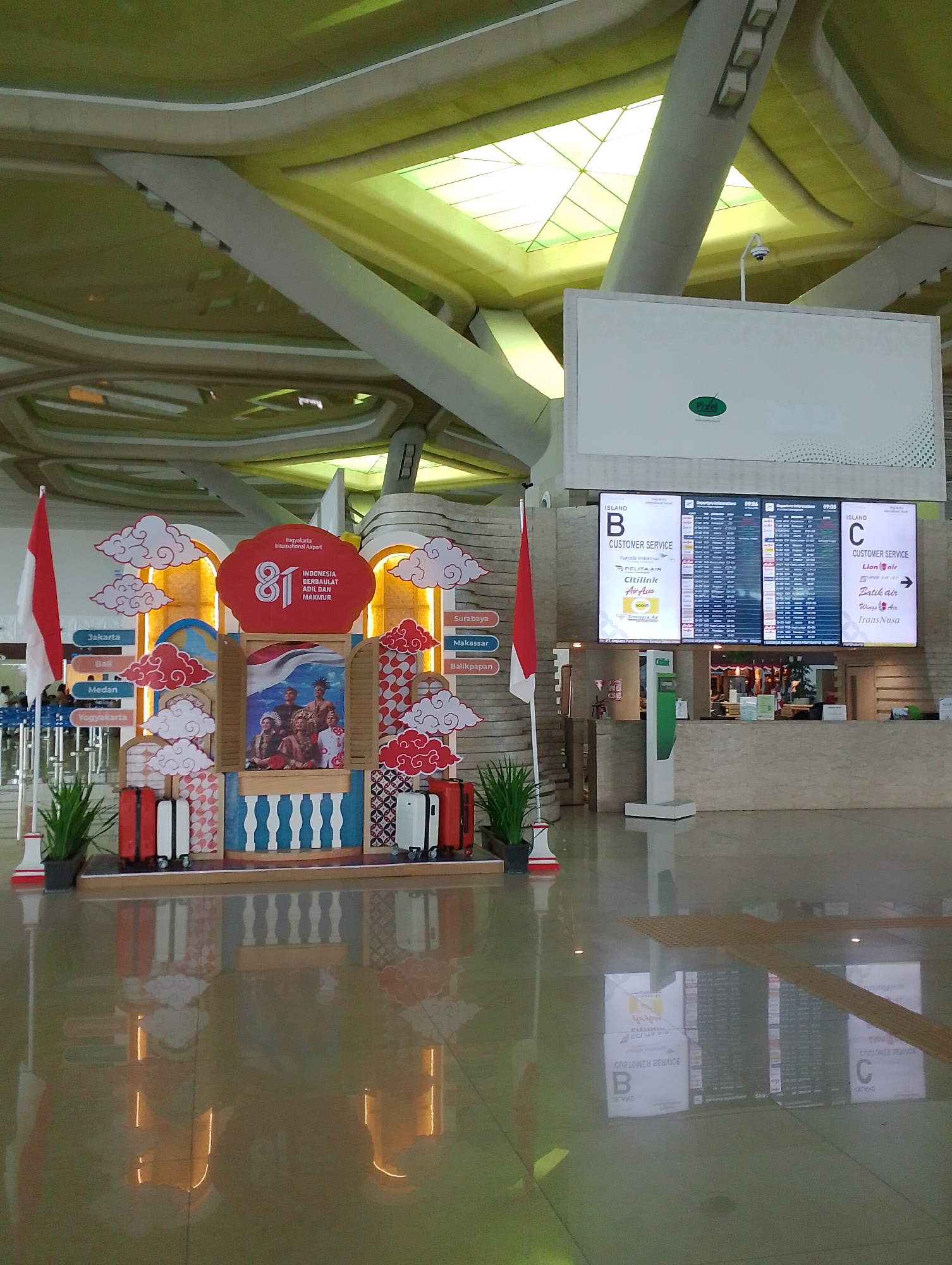 The image depicts the interior of Yogyakarta International Airport in Glagah, Indonesia, a modern airport terminal with a high, curved ceiling and large digital displays. In the foreground, a festive display is set up, featuring the number "81" and an image of people in traditional Indonesian attire. This display is flanked by red and white suitcases, and potted plants. The floor is highly reflective, showing a mirror image of the display and the terminal's ceiling lights. In the background, two large digital flight information boards are visible, one labeled "ISLAND B" and the other "ISLAND C". These boards list flight departures and arrivals. A security camera is mounted on a white pole above the "ISLAND B" display. The overall impression is of a busy and well-maintained airport environment, with a celebratory decoration likely related to a national event.