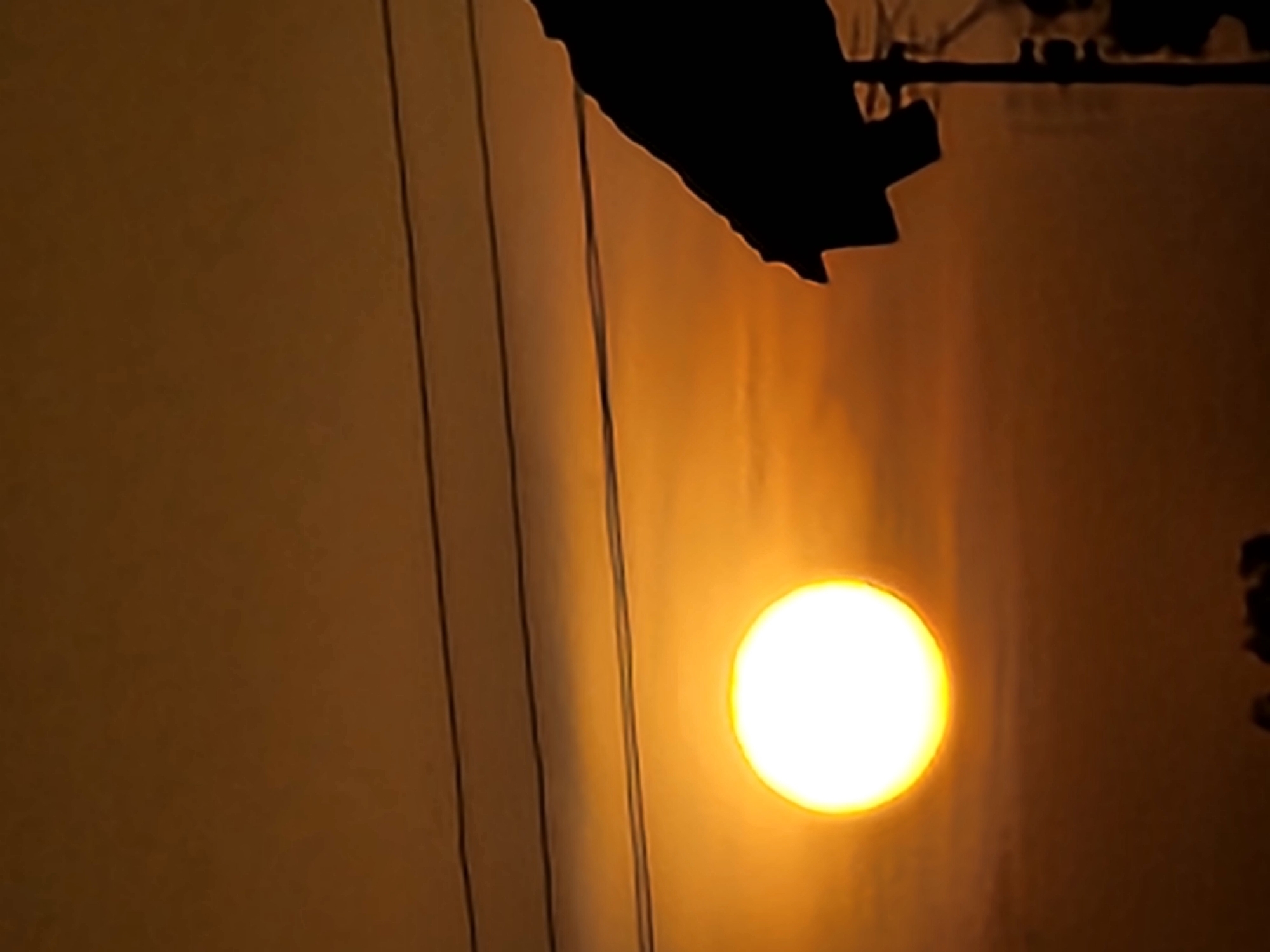 The image shows the sun, appearing as a bright yellow circle, partially obscured by what seems to be the edge of a building or structure. There are several vertical lines on the left side of the image, suggesting the presence of wires or cables. The color palette is dominated by yellowish-brown hues, indicating that it is either sunset or sunrise. It is difficult to determine the location without additional information. The image contains no discernible people or subjects.