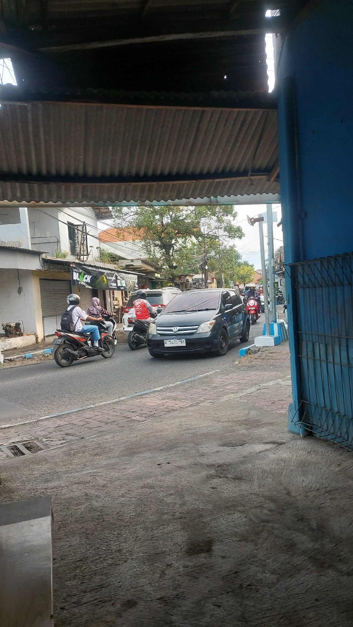 This is an eye-level, medium shot of a street scene in Sidoarjo, Indonesia, taken from under a covered structure. In the foreground, a rough concrete surface is visible. The main focus is a street with several vehicles and people. A dark gray hatchback car, possibly a Toyota Ist with license plate W 1435 NY, is moving in the right lane. To the left of the car, a person on a red motorcycle wearing a red vest and helmet is also in motion. Further left, another person on a black motorcycle, wearing a white shirt, blue jeans, and a white helmet with a backpack, is riding. Behind them, a person in a pink hijab on a scooter is also present.
On the left side of the street, a row of buildings is visible. The closest building has a closed metal shutter and a visible air conditioning unit. Adjacent to it, another building features a banner with the word "BLACK" on it. Trees with green foliage are scattered throughout the background, and utility poles and wires are also present. The street itself is paved with asphalt and has a lighter colored border. The scene appears to be a typical daytime street with moderate activity. The overhead structure in the foreground, made of corrugated metal, suggests an open-air shelter or awning.