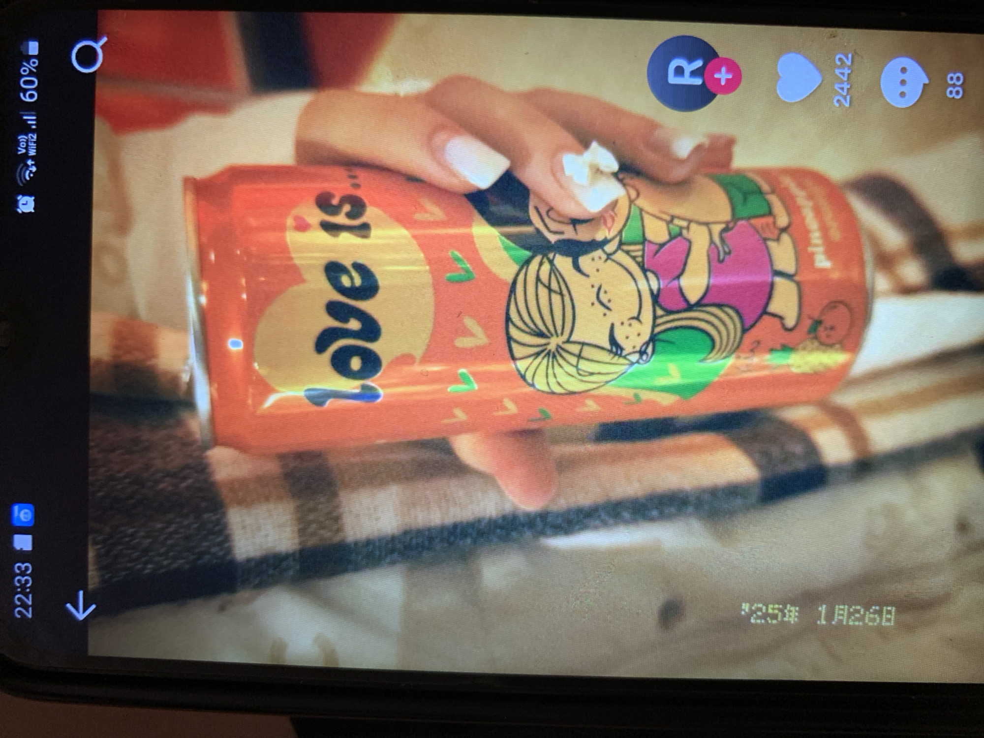 The image displays a close-up view of a person's hand holding an orange soda can with "love is..." text and cartoon characters on it. The background appears to be a blurred checkered pattern, possibly clothing or a blanket. The phone screen also shows a timestamp "22:33," network and battery status, and social media interface elements like a heart icon with "2442" likes and a comment icon with "88" comments. There is also a digital overlay of text vertically on the left side of the image, which appears to be "1254 1A26B". The scene is indoors, and the lighting suggests it was taken with a mobile phone camera in low light conditions. There is no information about the city of Lymanka, Ukraine, present in the image.