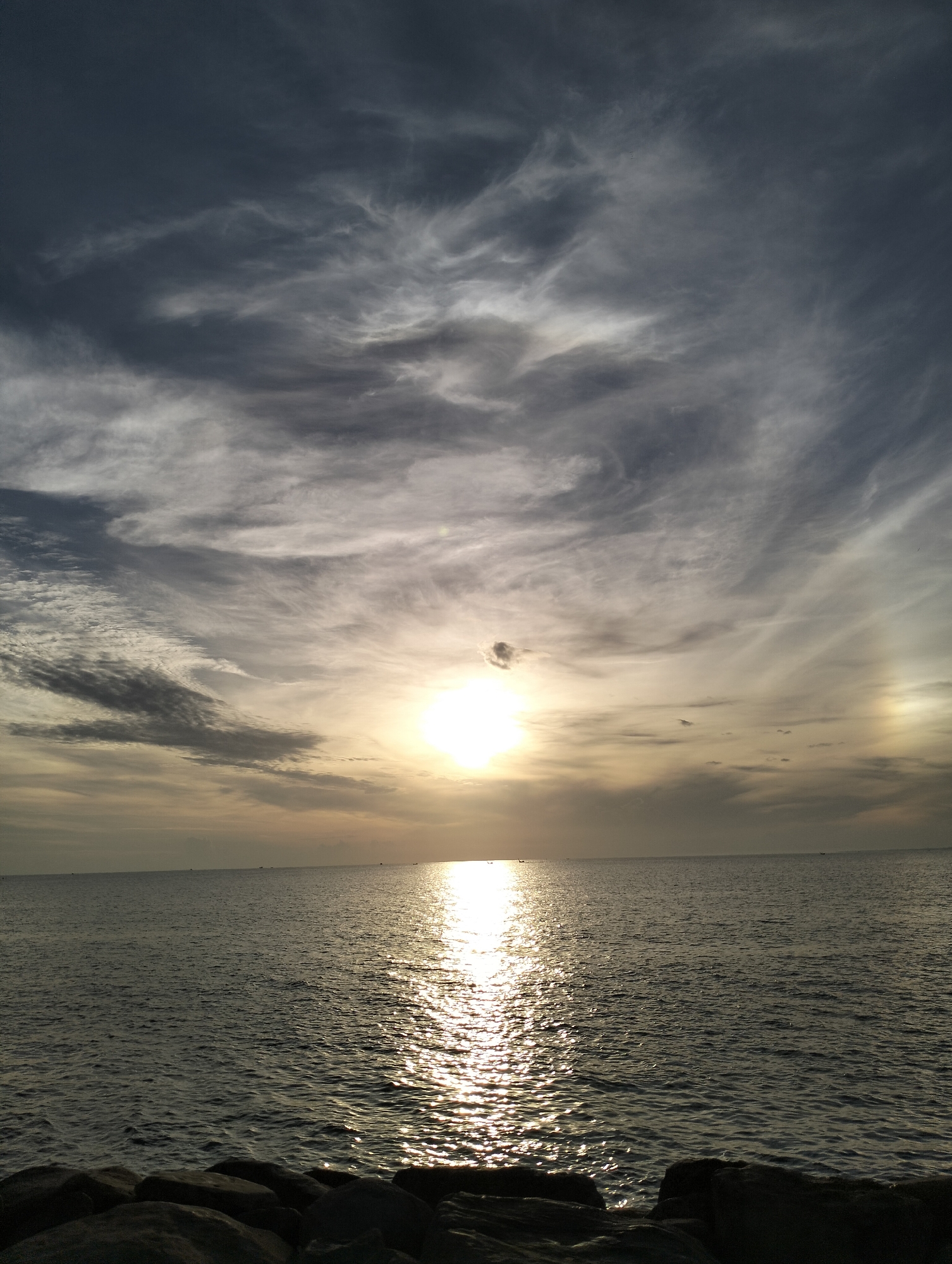 The image depicts a natural scene of the ocean at sunset, with a bright sun partially obscured by wispy clouds, casting a shimmering reflection on the water. The foreground features dark, silhouetted rocks, likely part of a breakwater or shoreline. The sky is a dramatic mix of dark gray and lighter, illuminated clouds, suggesting dynamic weather conditions. Several small, distant boats are visible on the horizon. The location is identified as the coast of Lhokseumawe, Indonesia.