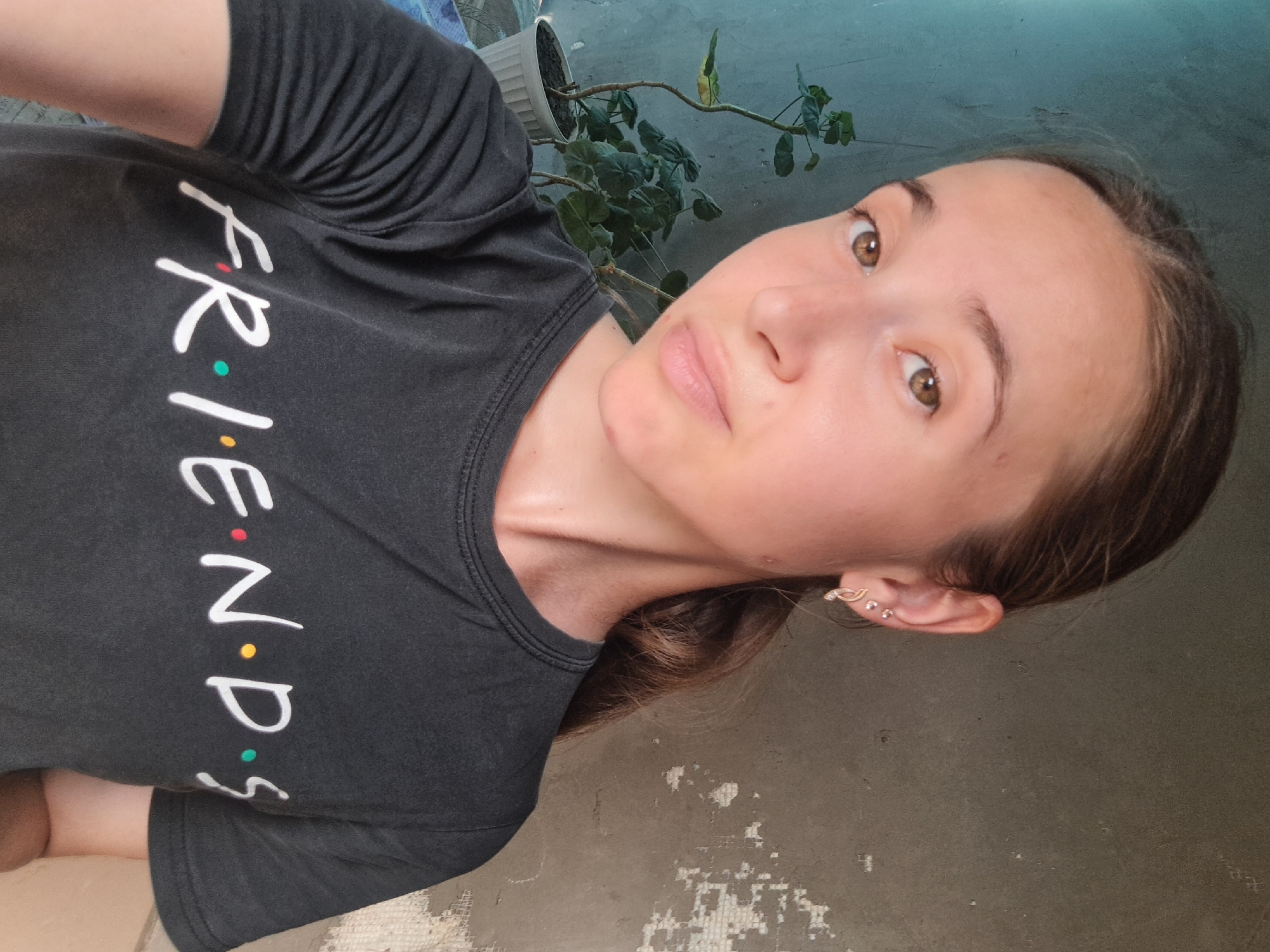 A young woman with light brown hair pulled back in a ponytail and brown eyes poses for a selfie. She is wearing a dark gray t-shirt with the word "FRIENDS" printed in white script with colored dots. She has on two small earrings in her left ear. Behind her, a potted plant with green leaves sits on a windowsill, and a textured gray wall is visible. The setting appears to be indoors, possibly in Vypasne, Ukraine.