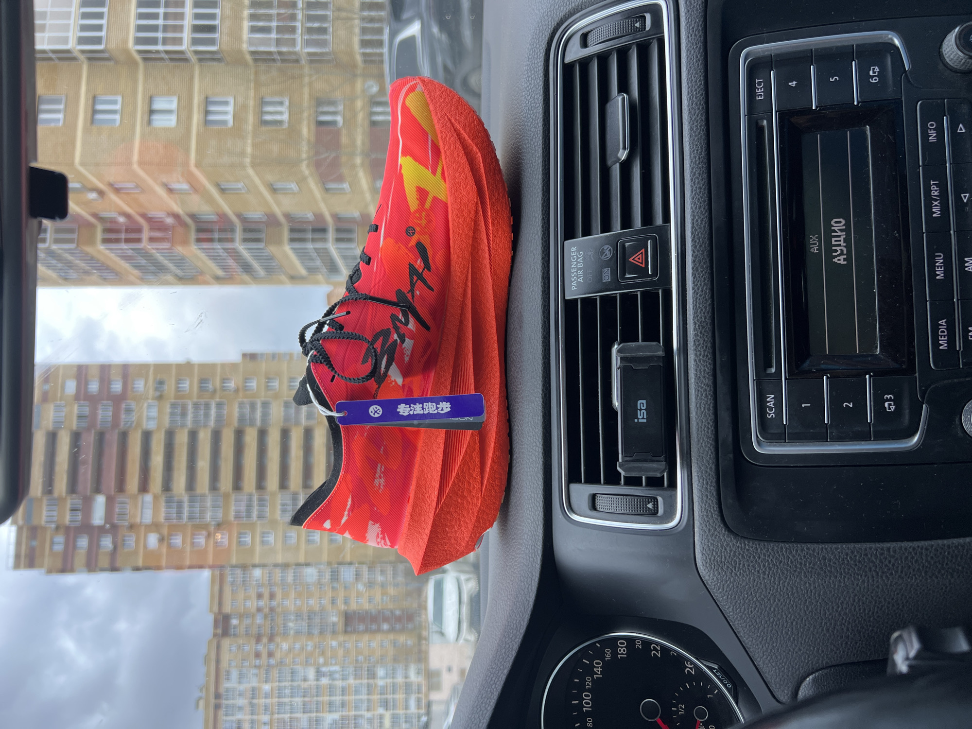 The image shows the interior of a car, specifically the dashboard area. A bright orange and red running shoe with black laces and a blue tag is placed on the dashboard, partially obscuring the passenger-side air vent. The car's radio and climate control panel are visible. Through the windshield, a multi-story building with a repeating pattern of windows can be seen. The building appears to be located in an urban area. Based on the car's dashboard and the building style visible through the windshield, the setting is Kazan, Russia.