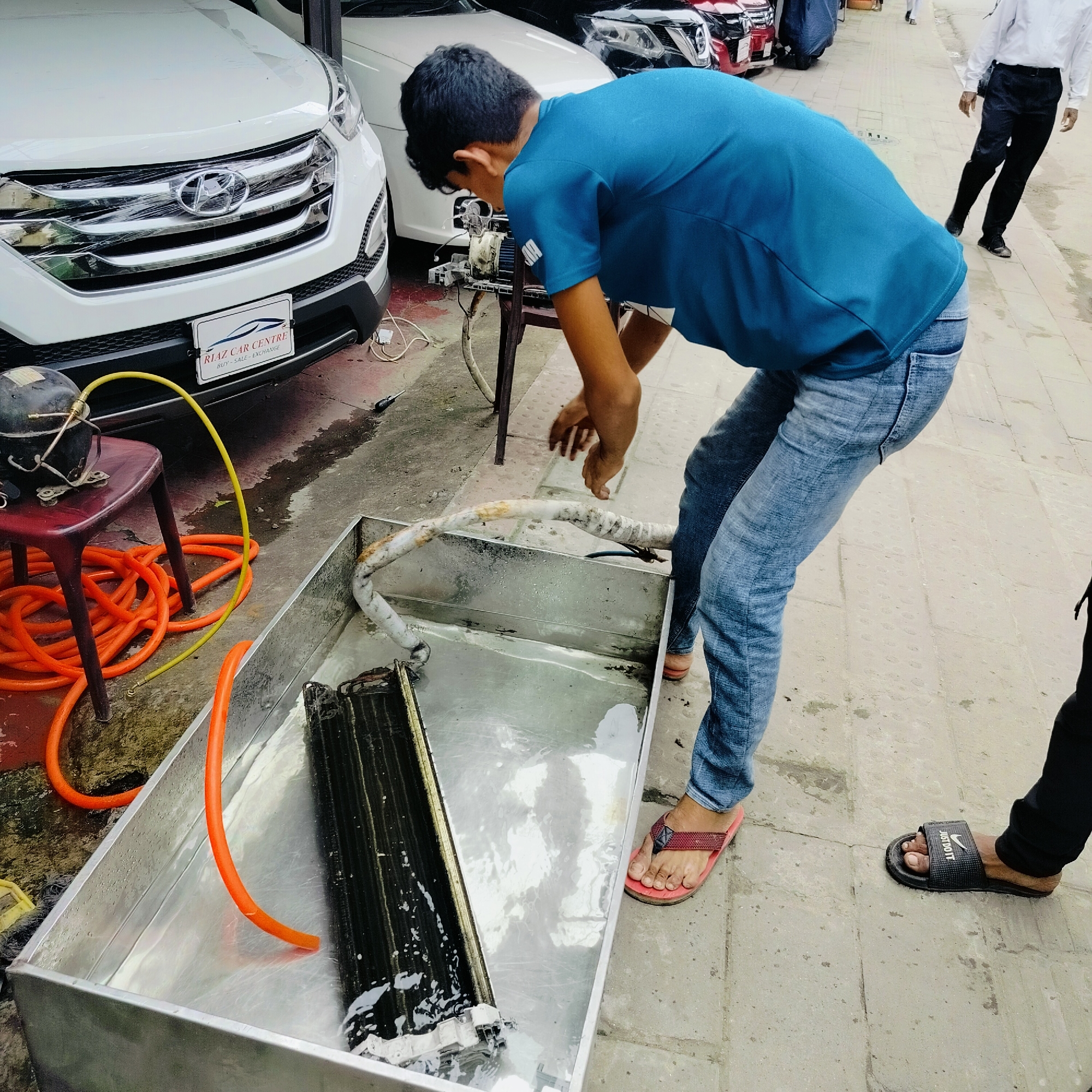 In an outdoor urban setting, a young man wearing a blue t-shirt and blue jeans is engaged in cleaning an air conditioner's condenser unit. He is bent over a large, rectangular metal basin filled with water and the condenser unit, which appears to be partially submerged. The condenser unit is black and has fins. He is holding a hose with his right hand, and a yellow hose is connected to a black compressor unit that sits on a red stool to the left of the basin. Several orange hoses are coiled on the ground near the stool. The ground is paved with concrete and appears wet in places. In the background, several cars are parked, including a white Hyundai and a red car. A person in a white shirt and black pants is walking away from the scene on the right. The overall impression is one of maintenance work being performed outdoors, possibly at a car service center or a similar establishment.