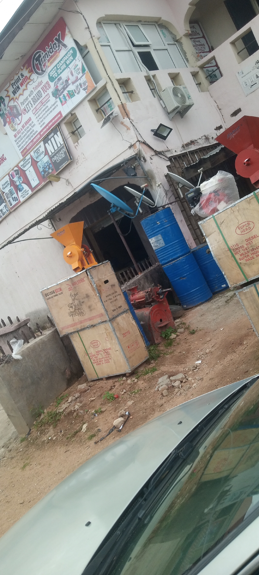 In Jalingo, Nigeria, an outdoor scene shows a building facade with various signs. Prominently displayed is a large red and white sign for "Trinidad," offering printing and branding services. Adjacent to it are smaller signs for "NIN General ICT Services" and "Enrollment Centre." Below these signs, stacked wooden crates and blue industrial barrels are visible. An orange agricultural machine, possibly a grinder, sits atop one of the crates. A silver car's windshield and hood occupy the foreground, angled upwards. The environment appears to be a commercial area with some overgrowth of weeds and scattered debris on the dusty ground. An air conditioning unit is mounted on the building's exterior.