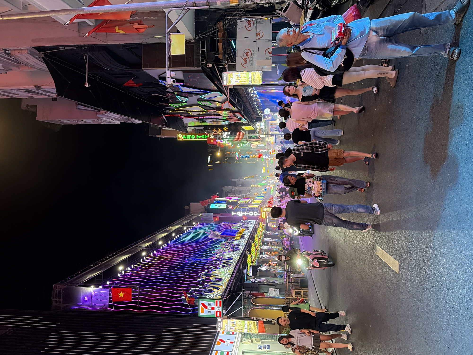 The image captures a bustling street scene at night, likely in Thủ Đức City, Vietnam, characterized by vibrant neon lights and a lively atmosphere. The street is paved and features a yellow dashed line down the center. On either side, buildings are adorned with colorful, illuminated signs and decorations, including wavy patterns of purple, pink, and blue lights on the left. Several flags, appearing to be Vietnamese flags, are visible.
Numerous people are walking along the street, some in small groups and others individually. They are dressed in casual attire, typical of a warm climate. Among the crowd, a person on a pink scooter is prominently featured, illuminating the road with its headlight. Other individuals are seen carrying items, such as bags and what appears to be a tray of goods held by an elderly man in the foreground. The buildings house various establishments, indicated by signs for "7 ELEVEN," "TATTOO," and other commercial entities. The overall environment suggests a commercial or entertainment district, with an urban setting at nighttime.