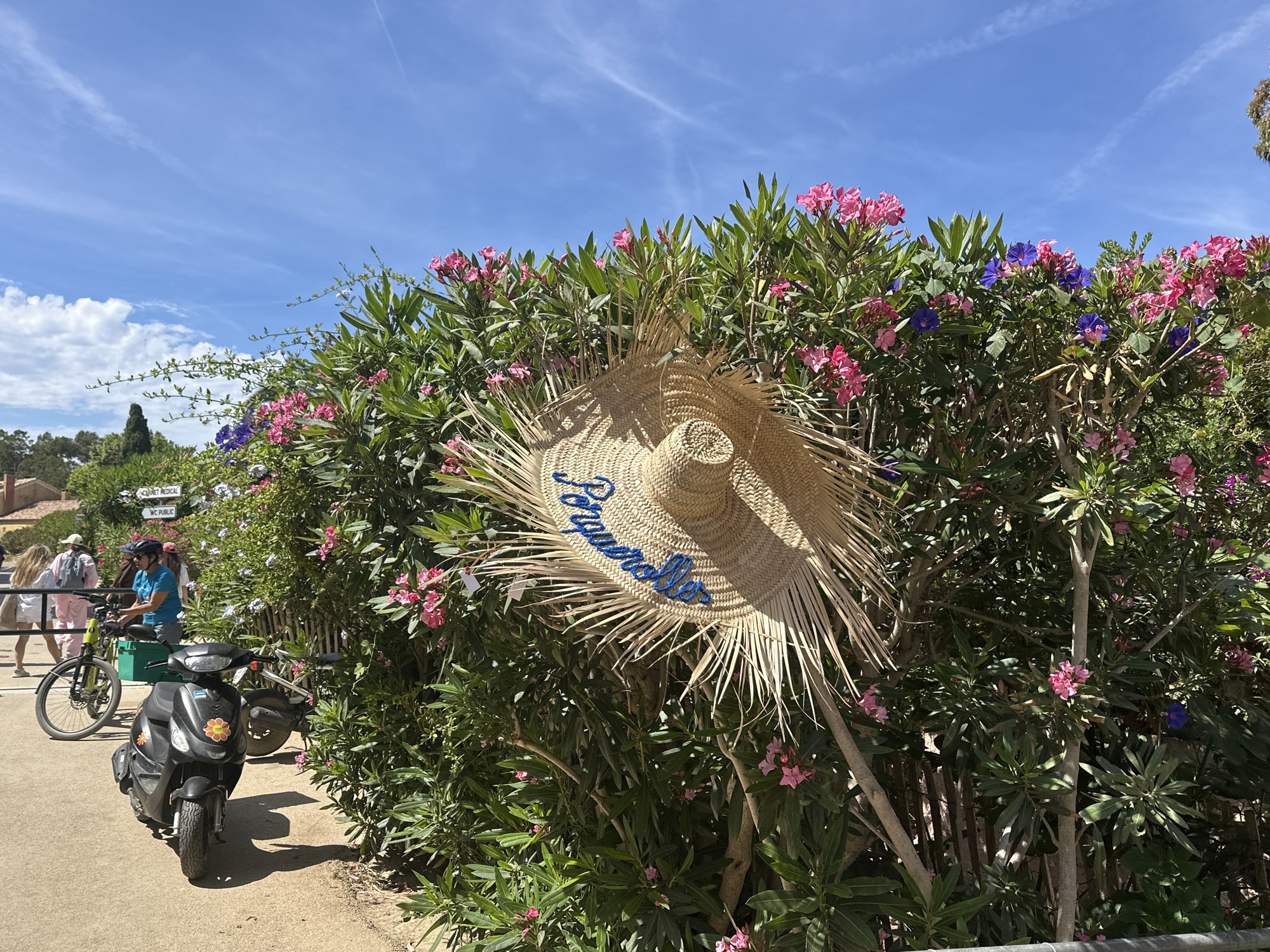 The image depicts a sunny outdoor scene with a large straw hat hanging on a hedge of pink and purple flowers. The hat has blue embroidered lettering that appears to say "Jacqueline". In the background, several people are visible, some walking and one on a bicycle. A black scooter with flower decals is parked in the foreground. Signs are visible on the hedge, one reading "Cabinet Medical" and another "WC PUBLIC", suggesting the location is a public area. The environment is lush and green, with a clear blue sky and some clouds. The scene is likely in a Mediterranean setting, given the vegetation and the overall ambiance, and possibly within Hyères, France, a city known for its coastal beauty and flora.