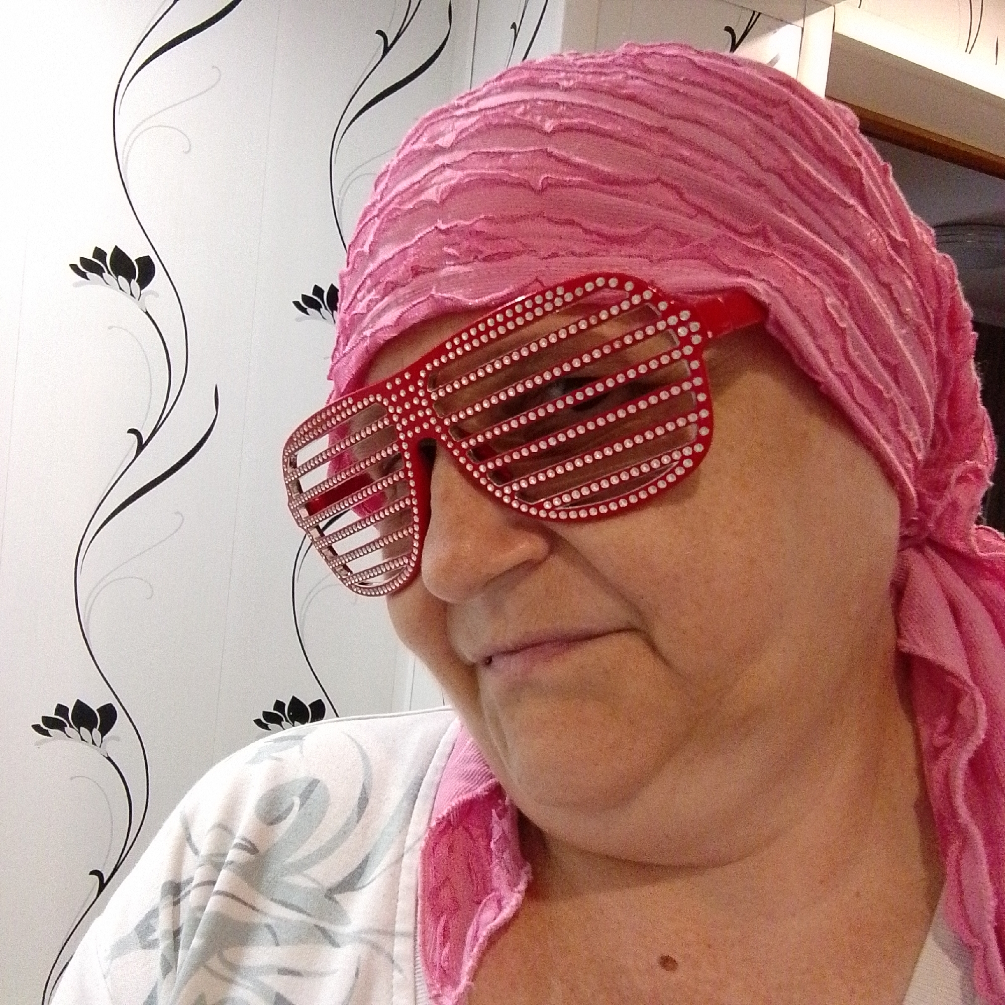 A person wearing a pink headscarf and red shutter shades is pictured in a close-up, indoor shot. The person's face is turned to the left and slightly downward, with their mouth partially open. The background features white wallpaper with a black floral pattern. The lighting appears to be artificial, creating a slightly warm tone. No specific location in Prokopyevsk, Russia is identifiable.