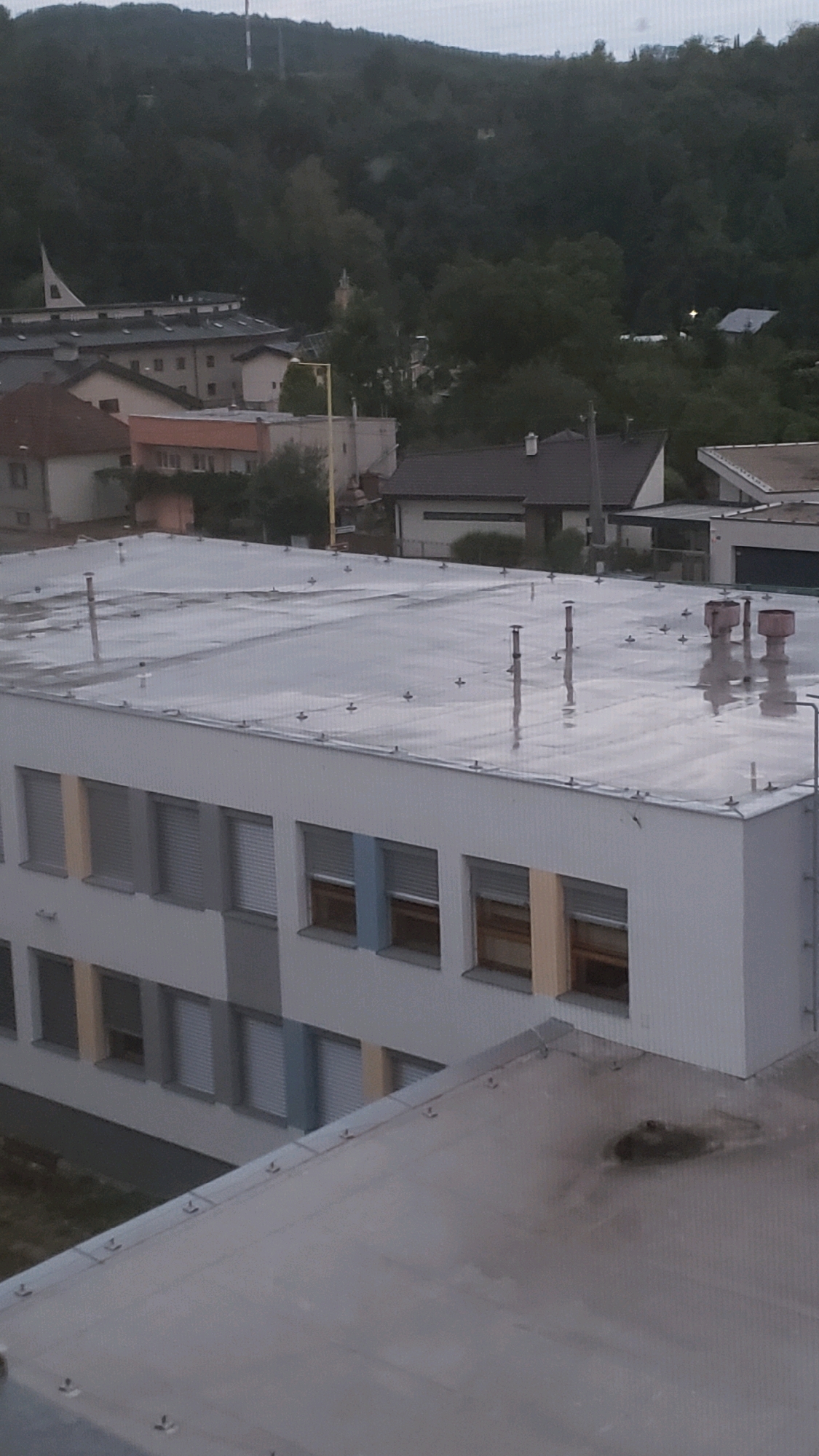 The image shows a view of a building in Prešov, Slovakia, with a hilly, tree-covered background. The foreground features a white, multi-story building with rows of windows. The roof of this building is flat and appears wet, with several vents or pipes protruding from it. In the middle ground, there are other buildings with pitched roofs, and a yellow pole with a light fixture is visible. The background consists of a dense forest covering the hills, with a visible communication tower or pylon on one of the peaks. The sky is overcast, suggesting a cloudy or rainy day. The overall scene is one of an urban or suburban environment surrounded by nature.