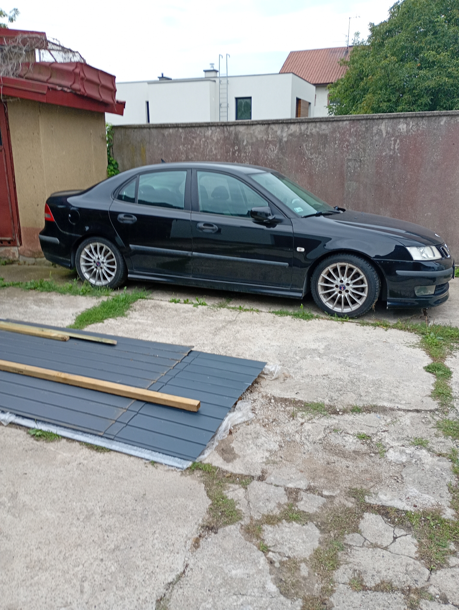 The image depicts a black Saab sedan parked on a cracked concrete driveway. To the left of the car, a section of a building with a red roof and beige siding is visible. In the background, a weathered concrete wall separates the driveway from modern buildings and a lush green tree. In the foreground, dark gray metal sheets and wooden planks are laid out on the concrete, suggesting ongoing construction or renovation work. The scene is outdoors, with an overcast sky indicating potentially inclement weather. The location is identified as Białystok, Poland.