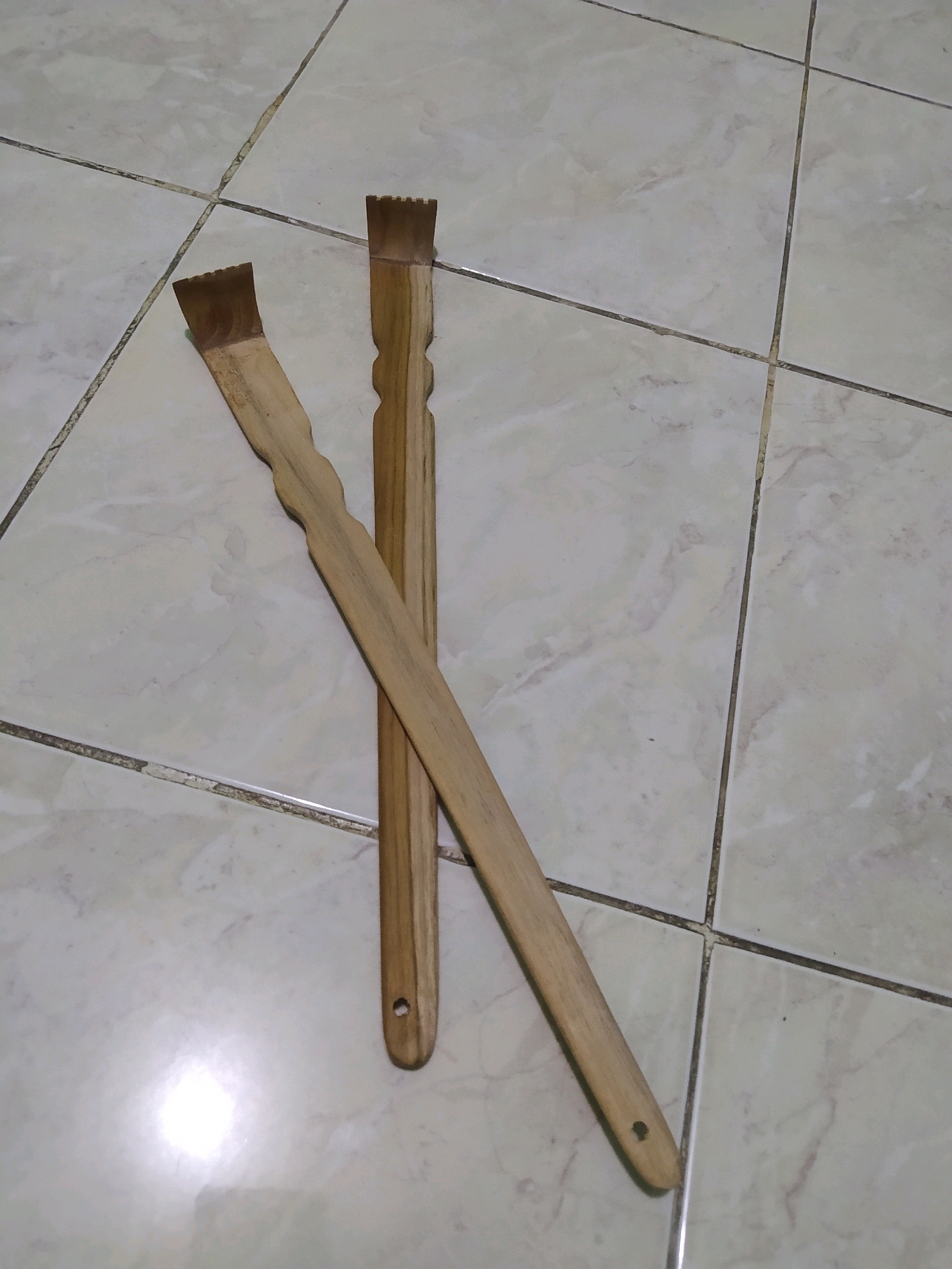 Two wooden back scratchers are placed on a light-colored tiled floor with dark grout lines. The scratchers are positioned diagonally across the frame, one on top of the other. The scratcher on top is slightly shorter and wider at the scratching end, which has a serrated edge. The scratcher underneath is longer and thinner, also with a serrated scratching end, and has a carved, wavy section in the middle of its handle. Both handles have a small hole near the end. The tiles appear to be square and arranged in a grid pattern. The lighting suggests an indoor environment, with a bright reflection visible on the floor. The image is taken from a slightly elevated perspective.