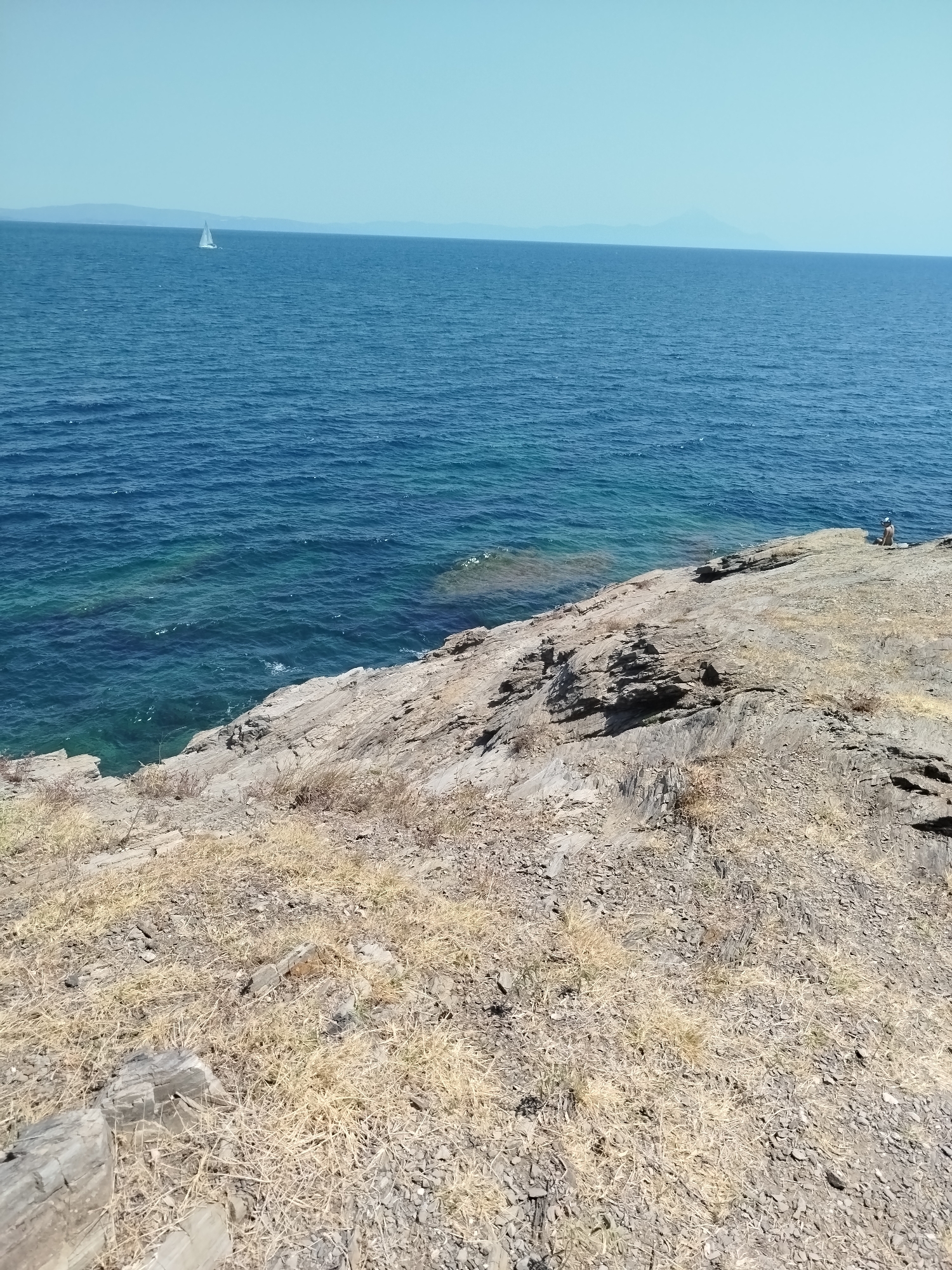 A wide-angle, eye-level shot captures a rocky coastline meeting the deep blue Aegean Sea under a clear, light blue sky. In the distance, a white sailboat is visible on the horizon. The foreground is dominated by dry, yellowish grass and scattered rocks. The middle ground consists of a rocky outcrop sloping down to the water. In the far right middle ground, a single person is seated on the rocks facing the sea. The water near the shore is a lighter, more translucent turquoise, gradually deepening to a darker blue further out. The landmasses of distant islands or the mainland are faintly visible on the horizon. The scene appears to be a natural, undeveloped coastal area within Sithonia Municipal Unit, Greece, likely during daylight hours given the brightness and clear sky.