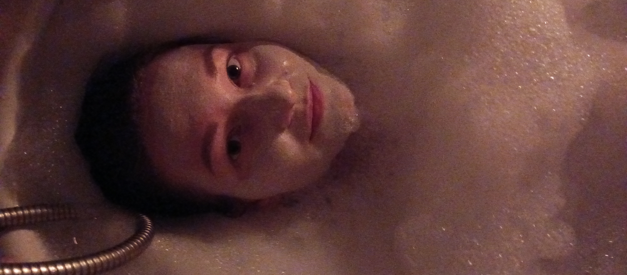 The image presents a close-up of a person's face and upper body partially submerged in a bubble bath. The individual is looking towards the viewer with a slight, content smile. Their face is partially obscured by white foam, particularly around the cheeks and chin. Their eyes are open, and lips are visible. The scene is dimly lit with warm, soft lighting, creating an intimate atmosphere. A metallic, flexible hose, possibly from a shower, is visible in the lower left corner of the frame. The surroundings are filled with abundant white bubbles, indicating a relaxing bath. No text is visible in the image.