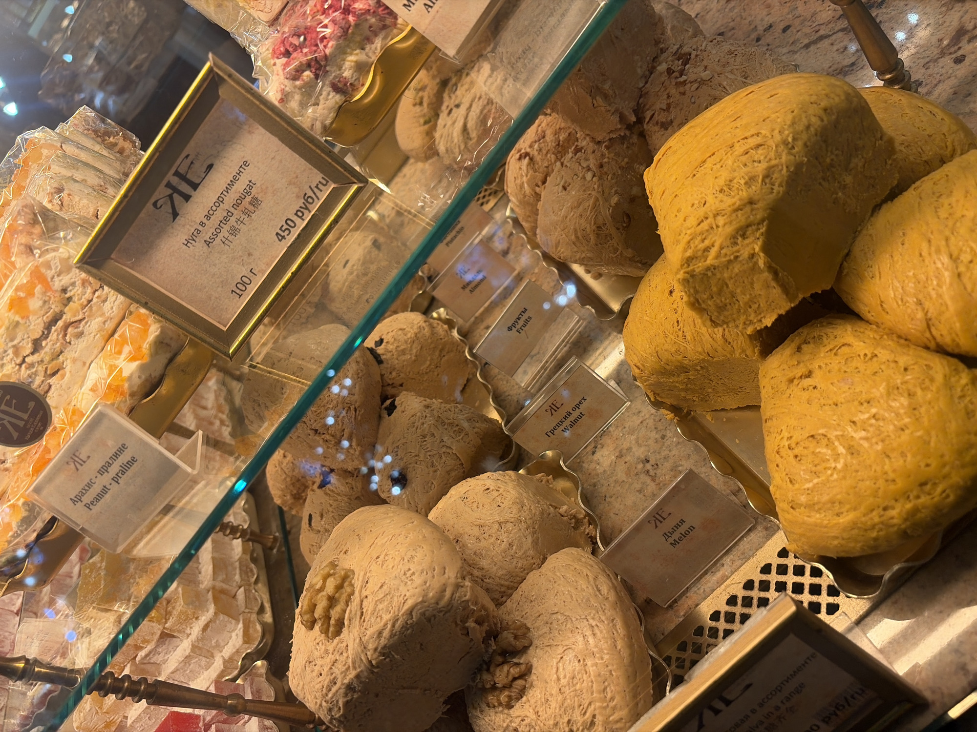 This is a close-up, eye-level shot of a display case filled with various sweets, likely a confectionery or bakery, in Saint Petersburg, Russia. The primary subjects are an assortment of halva and nougat products. The halva pieces are large, irregular blocks, some golden yellow and others a lighter, off-white color, with some containing visible nuts. The nougat appears to be sliced into smaller, rectangular pieces, some with visible fruit or nut inclusions.
Several small labels with text are visible throughout the display case, providing product names and prices in Russian and English. Examples include "Assorted nougat," "Peanut - praline," "Walnut," "Melon," "Pistachio," "Almond," and "Fruits." Prices such as "450 руб/ru" (450 rubles per item/unit) are also present. The display case is made of glass, with metal handles visible on some trays, suggesting a well-maintained shop environment. The lighting appears to be artificial, likely from overhead shop lights, creating some reflections on the glass. The overall impression is one of a tempting display of sweet treats, inviting customers to explore the variety.