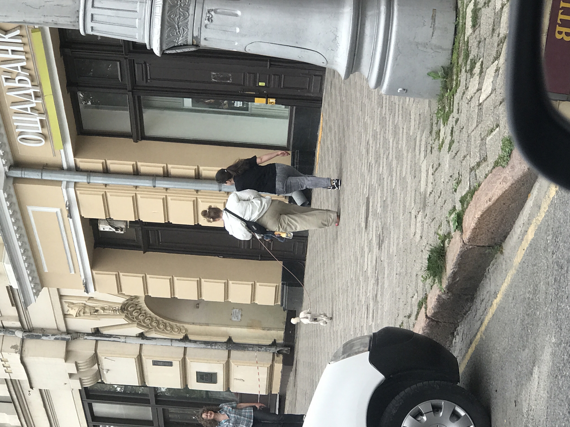 The image depicts a street scene in Kharkiv, Ukraine, with a building facade that includes the signage "ОЩАДБАНК" (Oschadbank). A woman with curly hair, wearing a plaid shirt and dark pants, stands on the left side of the frame near a red and white rope barrier. In the middle ground, two women walk away from the camera. One is wearing a white top and beige pants, and the other a black t-shirt and grey pants. A small white dog on a leash walks ahead of them, heading towards the right side of the image. A white car is partially visible in the foreground on the left, with its front bumper and wheel showing. The ground is paved with cobblestones, and a curb with some sparse grass is visible. A large, ornate lamp post stands on the right side of the frame. The overall scene suggests an urban environment on a cloudy day.