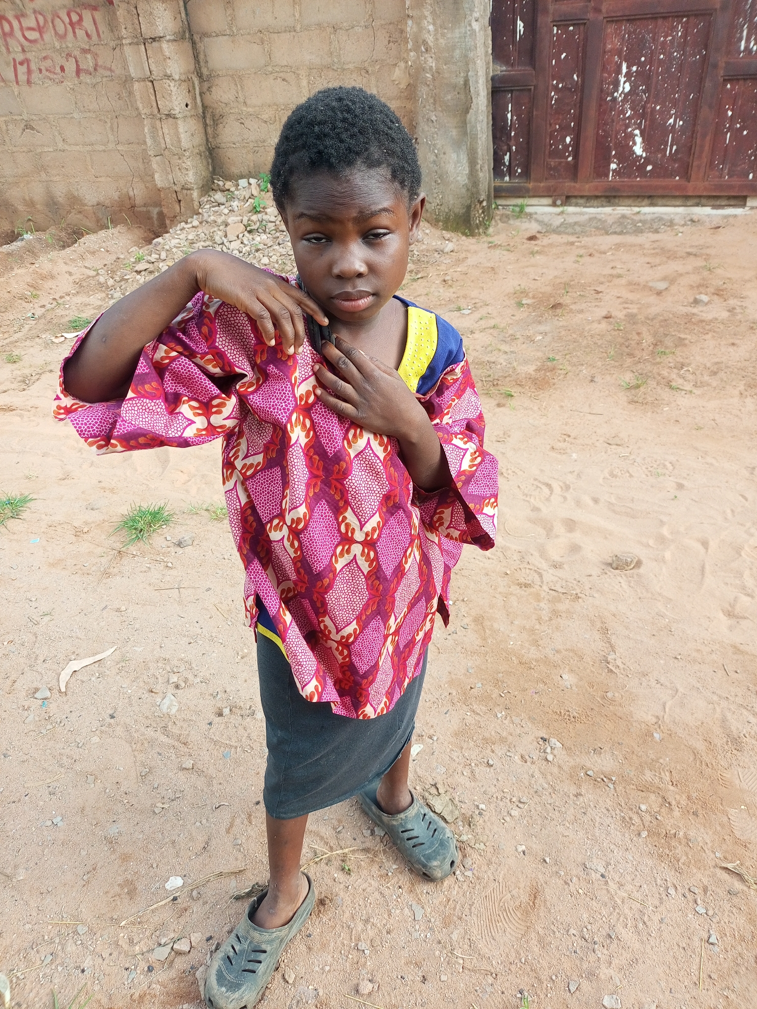 A young child, likely a girl, stands outdoors in an urban setting, possibly in Tempure, Nigeria. The child is wearing a vibrant, patterned shirt with wide sleeves and a dark skirt. She has dark hair and her gaze is directed slightly away from the camera. Her left arm is bent, and her hands are positioned near her chest, with the left hand resting on her right forearm. The child is barefoot and wearing dark, slip-on shoes that appear to be worn. The ground beneath her is a mix of sand and loose soil, with scattered small stones and dry grass. In the background, a rough, textured wall with graffiti is visible on the left. To the right, a dark, weathered wooden door with white paint splatters is partially visible. The overall impression is of a candid outdoor portrait, capturing a moment of the child's daily life in an arid, possibly developing, environment.