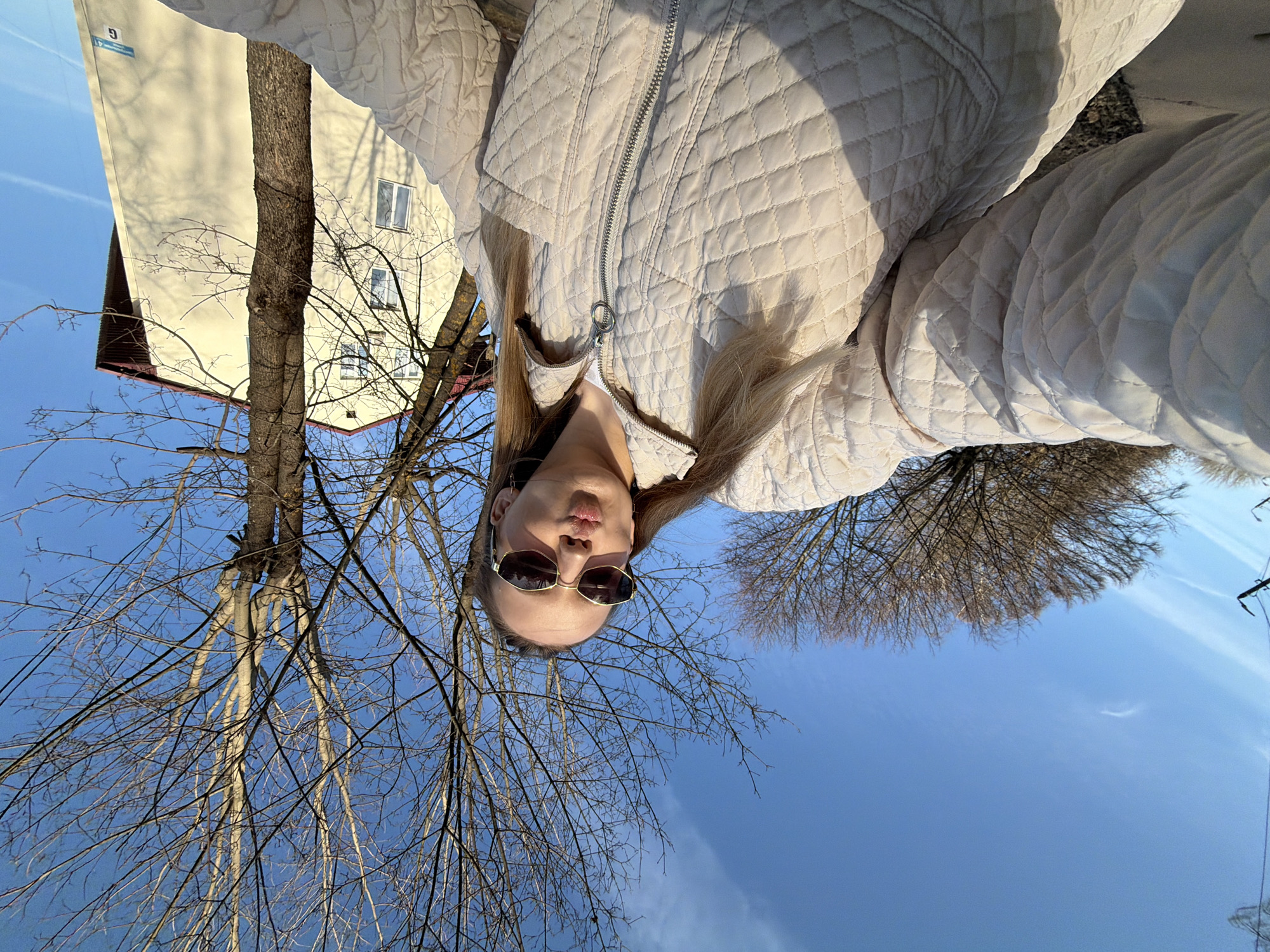 A woman wearing sunglasses and a light-colored quilted jacket is facing the camera in an upside-down perspective. Behind her, there is a light blue sky and bare trees with visible branches, suggesting it may be winter or early spring. A building with a cream-colored facade is partly visible on the left side of the frame. The image was taken in Reshetnikovo, Russia.