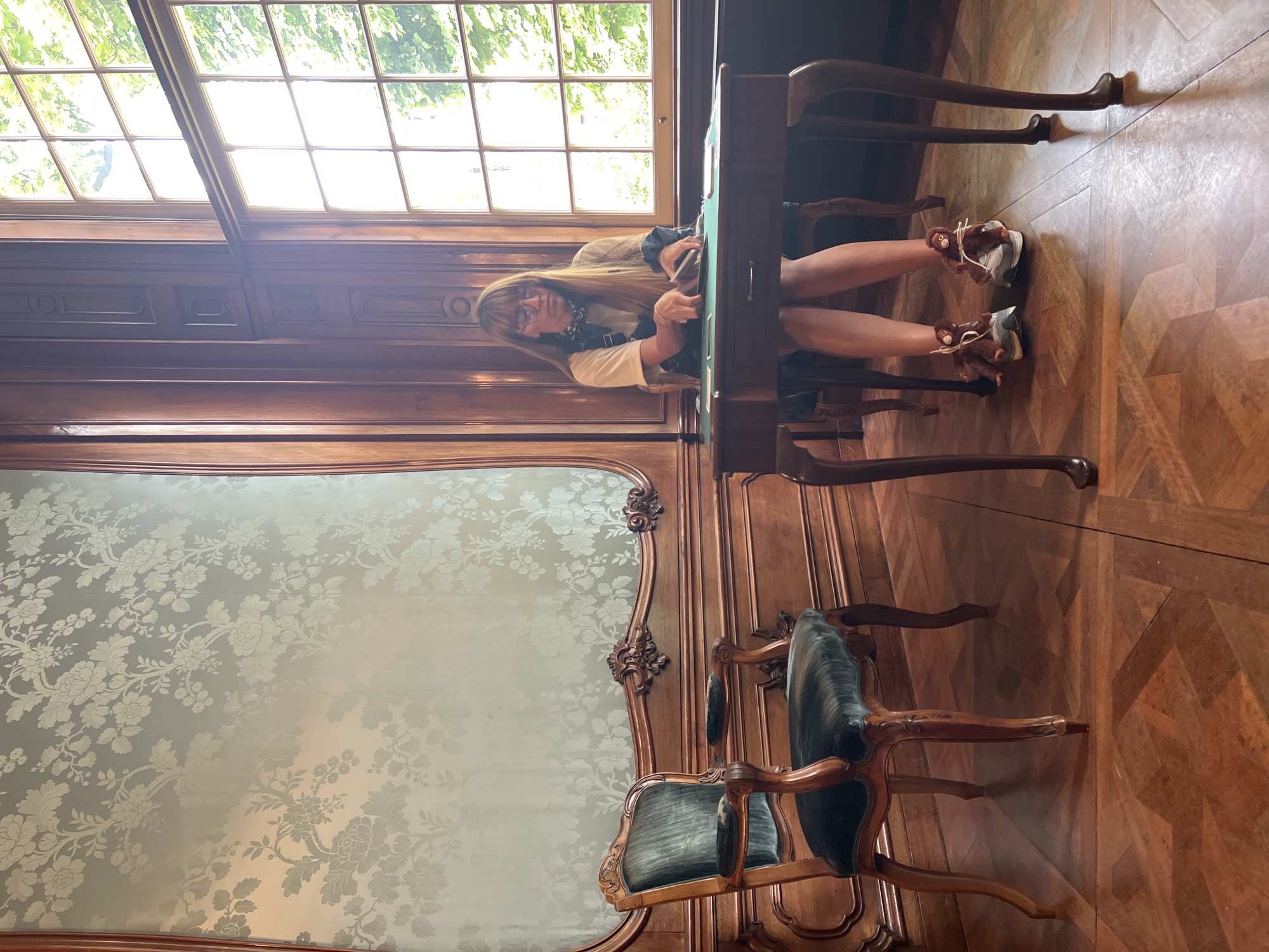 A woman with long brown hair and glasses sits at a dark wood table in a room with ornate wooden paneling and a patterned wallpaper. She is wearing a light-colored t-shirt, denim overalls, and sneakers adorned with stuffed monkey figures. Her hands are occupied with something on the table, which is covered with a green cloth. To her left is an antique armchair upholstered in dark green velvet. A large window to her right allows natural light to illuminate the scene, revealing trees outside. The floor is made of polished dark wood in a parquet pattern. The setting appears to be a historical interior, possibly a museum or grand residence, consistent with locations found in Amsterdam, Netherlands.