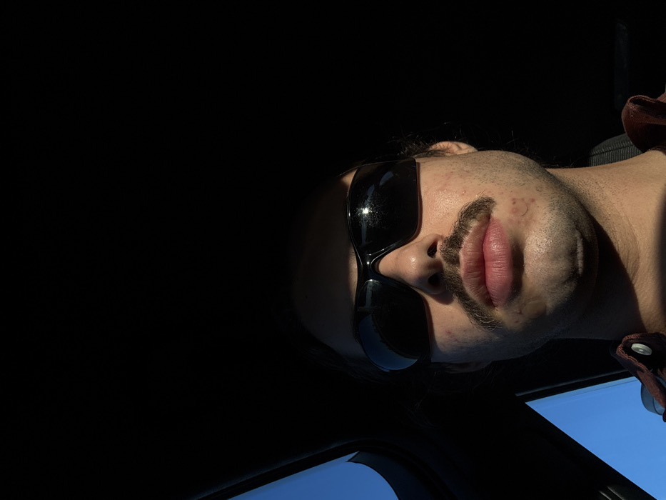 A male individual is depicted in a close-up, head-and-shoulders portrait, facing directly towards the camera within a vehicle in Lodi Vecchio, Italy. He wears dark, wraparound-style sunglasses and has a prominent dark mustache. Strong, direct sunlight illuminates the lower portion of his face, including his lips, chin, and mustache, while his forehead and the upper part of the vehicle's interior are in deep shadow. A circular, translucent patch is visible on the left side of his chin. He is wearing a reddish-brown collared shirt, partially visible at the bottom of the frame. A segment of clear blue sky is visible through a vehicle window on the left, contrasting with the dark interior.
