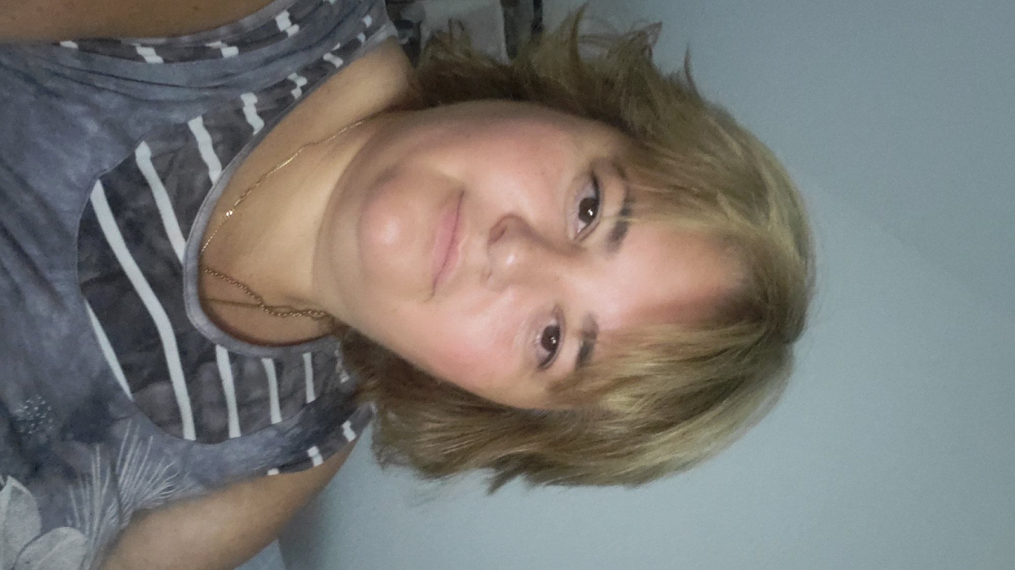 A medium shot shows a woman with shoulder-length blonde hair and bangs. She is wearing a grey and white striped tank top and a thin gold chain necklace. The background is a plain, light blue wall. She is looking slightly off-camera to her left, and a faint smile can be seen on her face. The image is taken from the chest up. There is no indication of a specific location in Sasbach, Germany, or any other city.