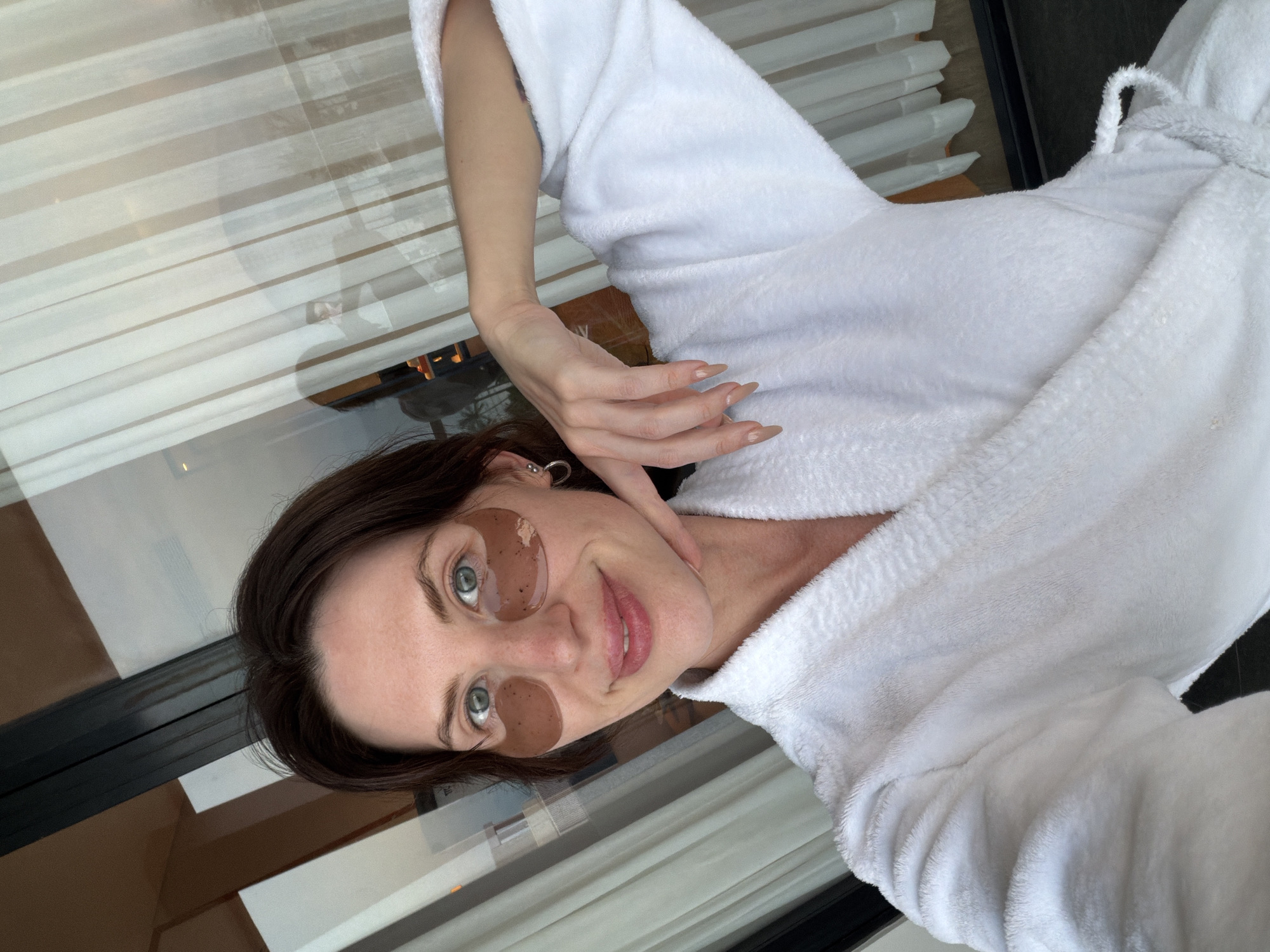 A light-skinned adult female with dark brown hair and light-colored eyes is depicted from the upper chest upwards, smiling directly at the viewer. She is wearing a white, plush bathrobe and has two brown, gel-like under-eye patches applied beneath her eyes. Her right hand, adorned with long, light-colored manicured nails and a silver hoop earring visible on her left ear, is raised with the index finger touching her chin. The background consists of a large glass window or door, through which reflections are visible. On the left side of the reflection, elements of an interior space, including a dark vertical frame and a lighter wall or wooden panel, can be seen. On the right side, a light-colored, vertically striped curtain is partially drawn, and reflections of a blurred outdoor scene with trees or foliage are present, along with a faint, dark silhouette of another person. The scene suggests an indoor setting or a balcony adjacent to an indoor space. The image does not contain visual cues to confirm its location in Dong Ninh Hoa Ward, Vietnam.
