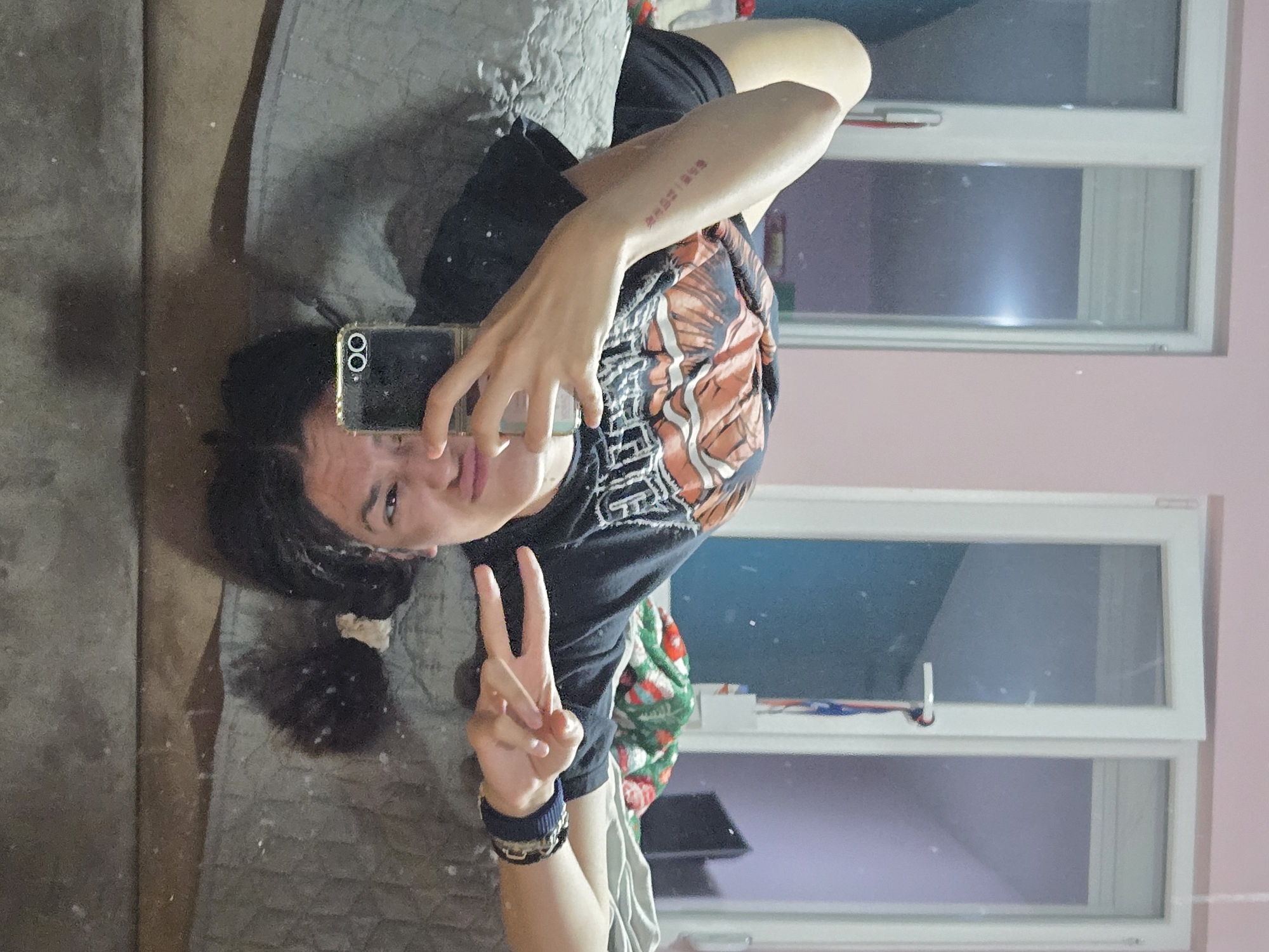 A young person with dark hair pulled back in a low ponytail is taking a selfie in a mirror. The individual is wearing a black t-shirt with an orange and white graphic design and is holding a black smartphone with multiple camera lenses. A peace sign is displayed with the hand not holding the phone. The arm visible in the mirror has a tattoo with Chinese characters. The background includes a gray bedspread, a pink wall, and a white-framed window with multiple panes, allowing light into the room. The scene appears to be indoors, possibly a bedroom. There are no indications of location in Pleven, Bulgaria.
