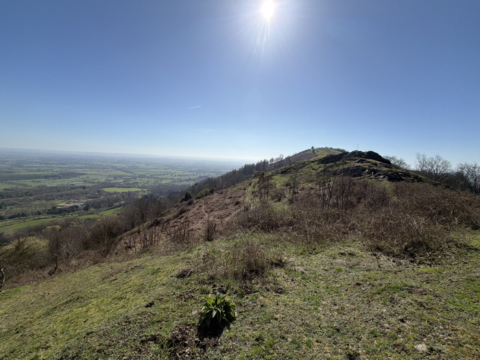 The image displays an elevated landscape scene within the Malvern Hills, United Kingdom, on a clear, sunny day with the sun visible in the upper-left quadrant. The foreground consists of a gently sloping area covered in patchy green grass and brown, dry vegetation, with a small cluster of bright green foliage centrally located at the bottom. The midground features a prominent hillside or ridge, partially covered with rocky outcrops near its crest, and slopes characterized by sparse deciduous trees, some bare, intermingled with green grass and brown undergrowth. A dirt path or trail is discernible traversing the hillside on the left, leading towards a faintly visible small marker or structure on a more distant, higher point of the ridge. The background extends into a vast, panoramic view of a valley and plains below, presenting a mosaic of green agricultural fields, darker wooded areas, and indistinct distant structures, all receding towards a hazy horizon. No human subjects, animals, or explicit actions are depicted.