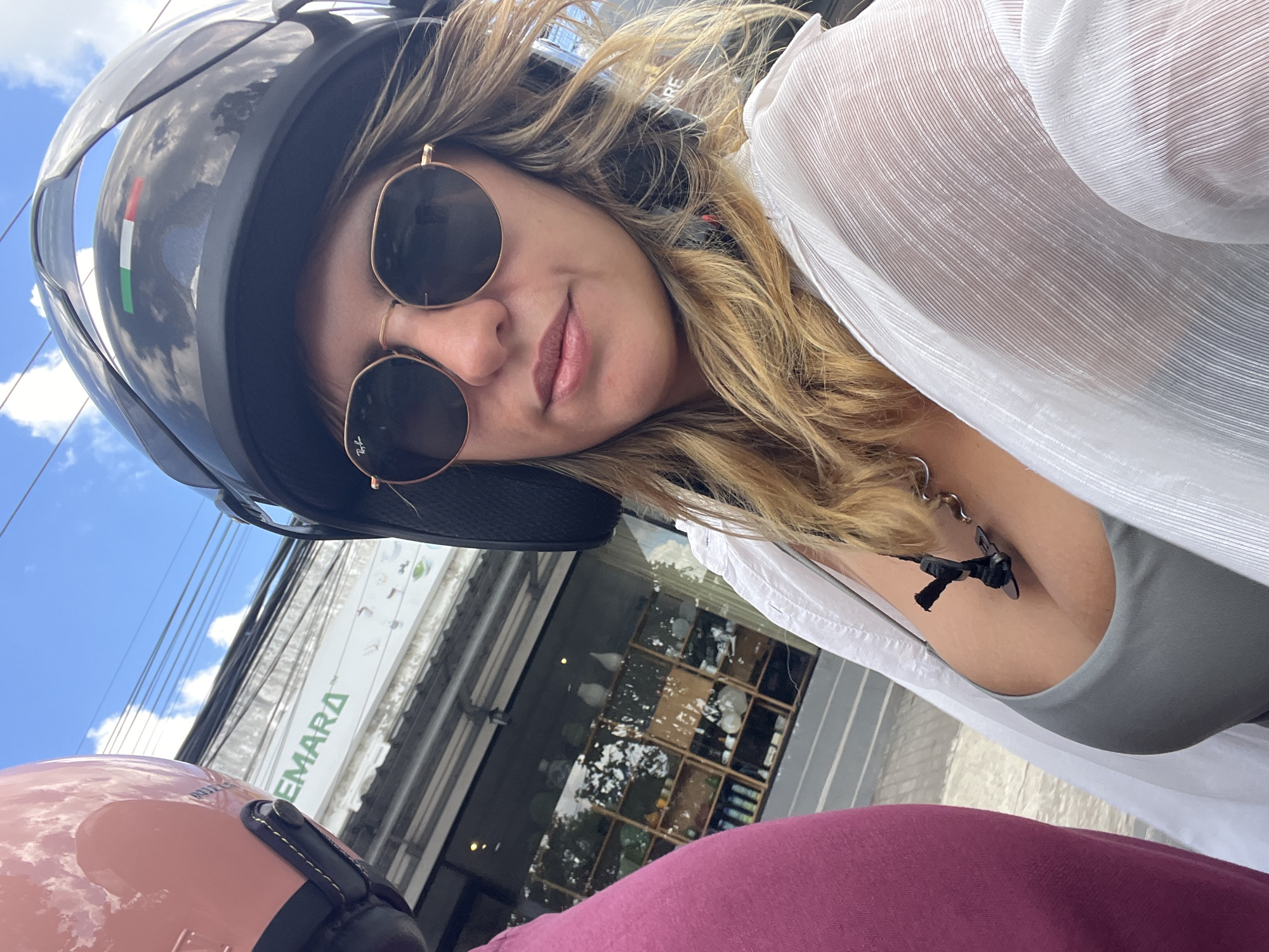 A close-up, slightly angled selfie shows two people on a motorcycle in front of a shop. The person in the foreground, wearing a black helmet with an Italian flag sticker and round sunglasses, is looking at the camera. They have blonde hair and are wearing a sheer white shirt over a gray tank top. A black lanyard with a clip hangs from their neck. To the left, the shoulder and head of another person wearing a pink helmet are visible. The shop behind them has a sign that reads "EMARA" in green letters, and the interior with shelves is visible through the window. The scene appears to be on a street in Mengwi, Indonesia, with a blue sky and power lines in the background.