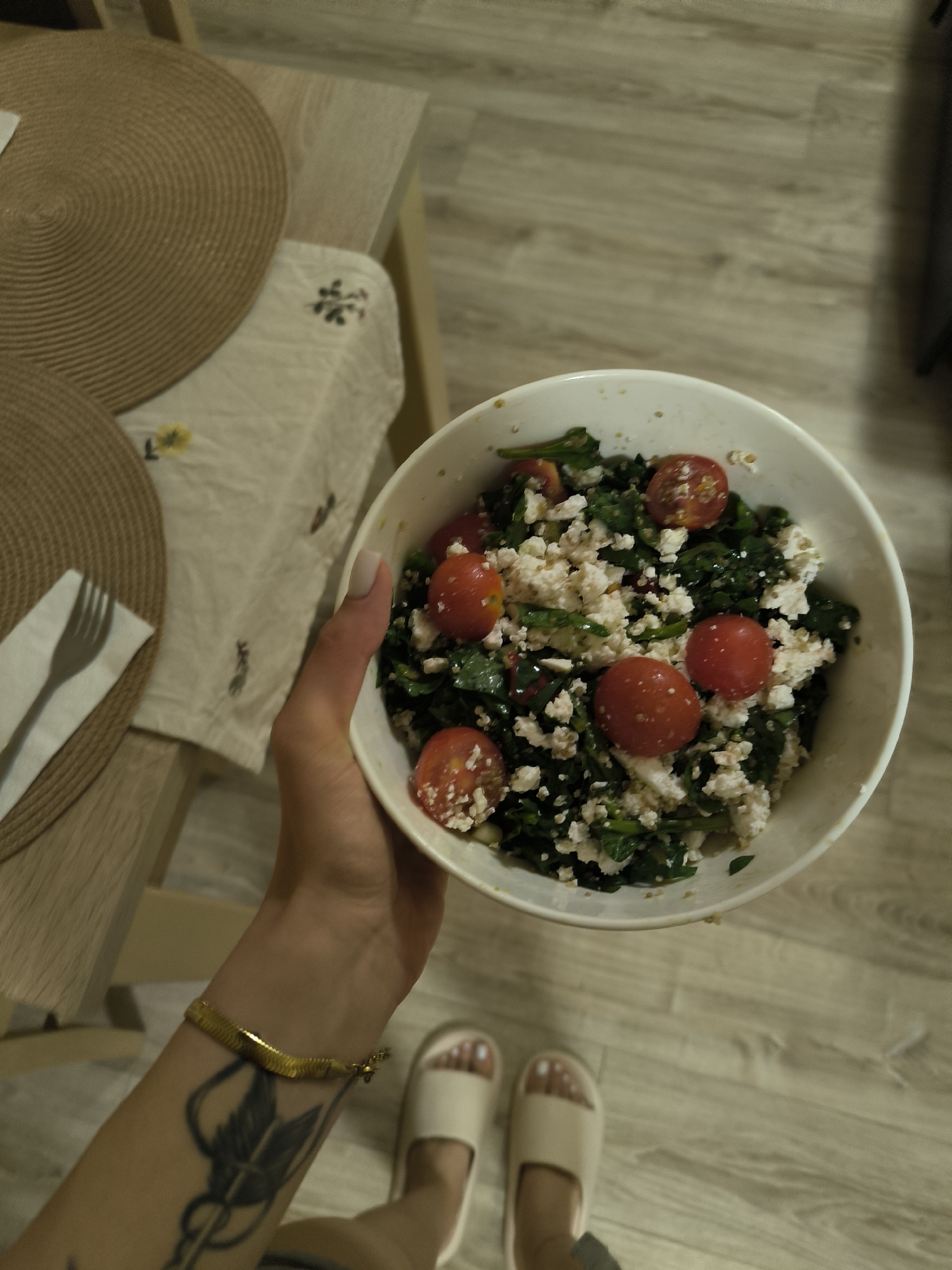 An overhead, slightly angled shot depicts a person holding a white bowl of salad over a light-toned wooden floor. The person's right forearm and hand are visible in the lower center, with a golden snake chain bracelet on the wrist and a dark tattoo resembling an arrow or spearhead on the inner forearm. The person's feet, clad in light beige slip-on sandals, are visible in the lower right, standing on the same wooden floor. The white bowl contains a salad composed of dark green leafy vegetables, several halved red cherry tomatoes, and a substantial amount of crumbled white cheese or curd, possibly with small grains like quinoa mixed in. To the left, a light wooden table is set with two circular woven placemats in a light brown hue. A light-colored fabric runner, featuring subtle yellow floral patterns and dark embroidered symbols, is draped over the table beneath the placemats. A folded white napkin with a silver-colored fork rests on the upper placemat, casting a distinct shadow. The background floor consists of light wooden planks. The lighting appears to be indoor artificial illumination.
