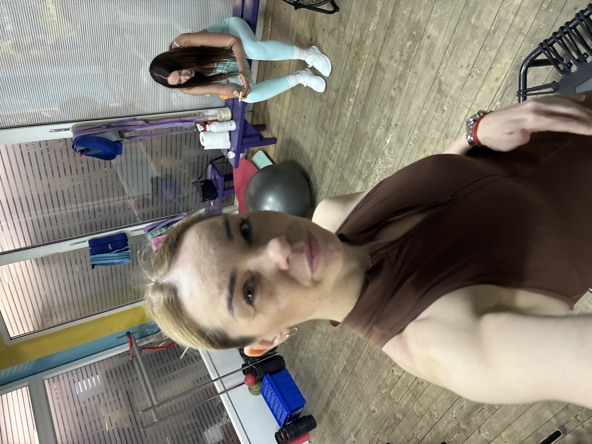 An interior shot, rotated 90 degrees clockwise, captures a selfie being taken by a woman in the foreground within what appears to be a fitness studio or gym. The main subject, positioned on the right side of the image (when viewed upright), has blonde or light brown hair and is wearing a dark brown sleeveless top. Her facial expression is neutral, and her left arm, adorned with a red and silver bracelet, is visible.
In the mid-ground, a second woman with long dark hair is seen bent over a purple wooden bench. She is wearing light blue leggings, a light-colored top, and white sneakers with white socks. She appears to be focused on items on the bench, possibly adjusting something or preparing for an activity. On the bench, a roll of white paper towels, a small white bottle, and various colorful resistance bands are visible. A blue bag or pouch hangs from the bench.
The setting features a light wooden floor and large windows with vertical frosted or striped patterns, suggesting an indoor space with natural light, though the time of day is not clearly discernable. Several pieces of gym equipment are scattered around, including a large black exercise ball, pink and green mats, blue storage bins, and black foam rollers (visible in cropped sections). Metal racks and bars for exercise equipment are also present. The walls near the windows are painted yellow and light blue in sections. There is no visible text in the image. The overall atmosphere suggests a casual workout environment.