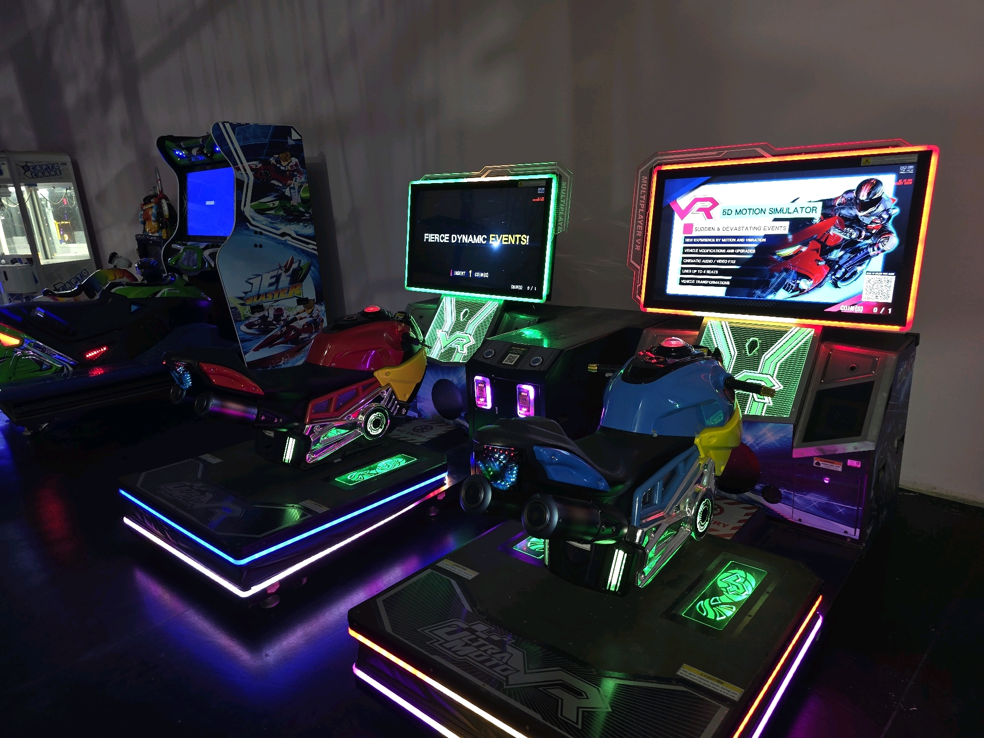 This is a medium shot, eye-level, indoor photograph of several arcade racing simulators. The simulators are themed as motorcycles and are set up in what appears to be an entertainment venue. The lighting is dim, with the primary illumination coming from the colorful LED lights on the simulators and their bases, which cast vibrant purple and blue hues on the dark floor.
On the left side of the image, a simulator with a red motorcycle seat is visible. Its base has green and white LED lighting. To its right, another simulator with a blue motorcycle seat is positioned. This one features a more prominent blue LED accent on its base, with green lights integrated into the platform.
In the background, to the left, a claw machine arcade game is partially visible. It is white with "GRABTO CRANE" branding. Further to the right, a tall, upright arcade cabinet with a screen displaying a blue background and what appears to be game graphics for "JET BLASTER" is present.
The screens of two of the motorcycle simulators are illuminated. The left simulator's screen displays "FIERCE DYNAMIC EVENTS!" in white text against a black background, with "INSERT 1 COIN(S)" at the bottom. The right simulator's screen shows "VR 5D MOTION SIMULATOR" prominently, along with text describing features like "SUDDEN & DEVASTATING EVENTS!", "NEW EXPERIENCE BY MOTION AND VIBRATION", "VEHICLE MODIFICATIONS AND UPGRADES", "CINEMATIC AUDIO / VIDEO FX", "LINKS UP TO 4 BEATS", and "VEHICLE TRANSFORMATIONS." The screen displays an image of a person in racing gear on a motorcycle.
The simulators are all empty, with no people present. The environment appears to be a commercial arcade or entertainment center, likely located in Bodrum, Turkey, given the context of the request. The floor is a dark, reflective surface.