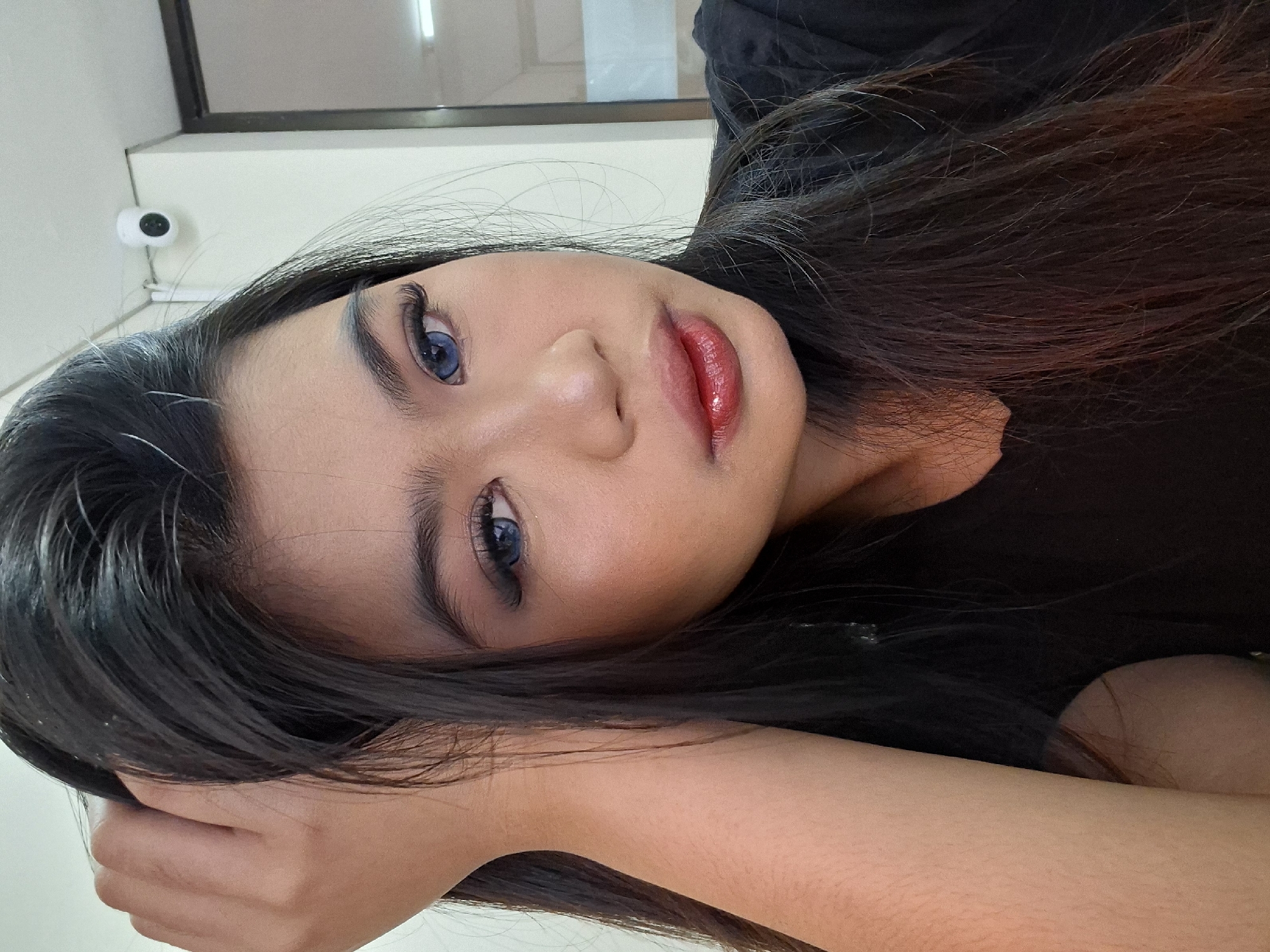 A young woman with long, dark hair, wearing blue contact lenses and heavy makeup, poses for a selfie indoors. Her right arm is raised, with her hand resting on her head, her fingers tangled in her hair. She is looking directly at the camera with a neutral expression. She is wearing a black shirt. A security camera is visible on the wall in the background. The location is an indoor space, possibly an office or a public building. There is no indication of the city of Kuta, Indonesia in the image.