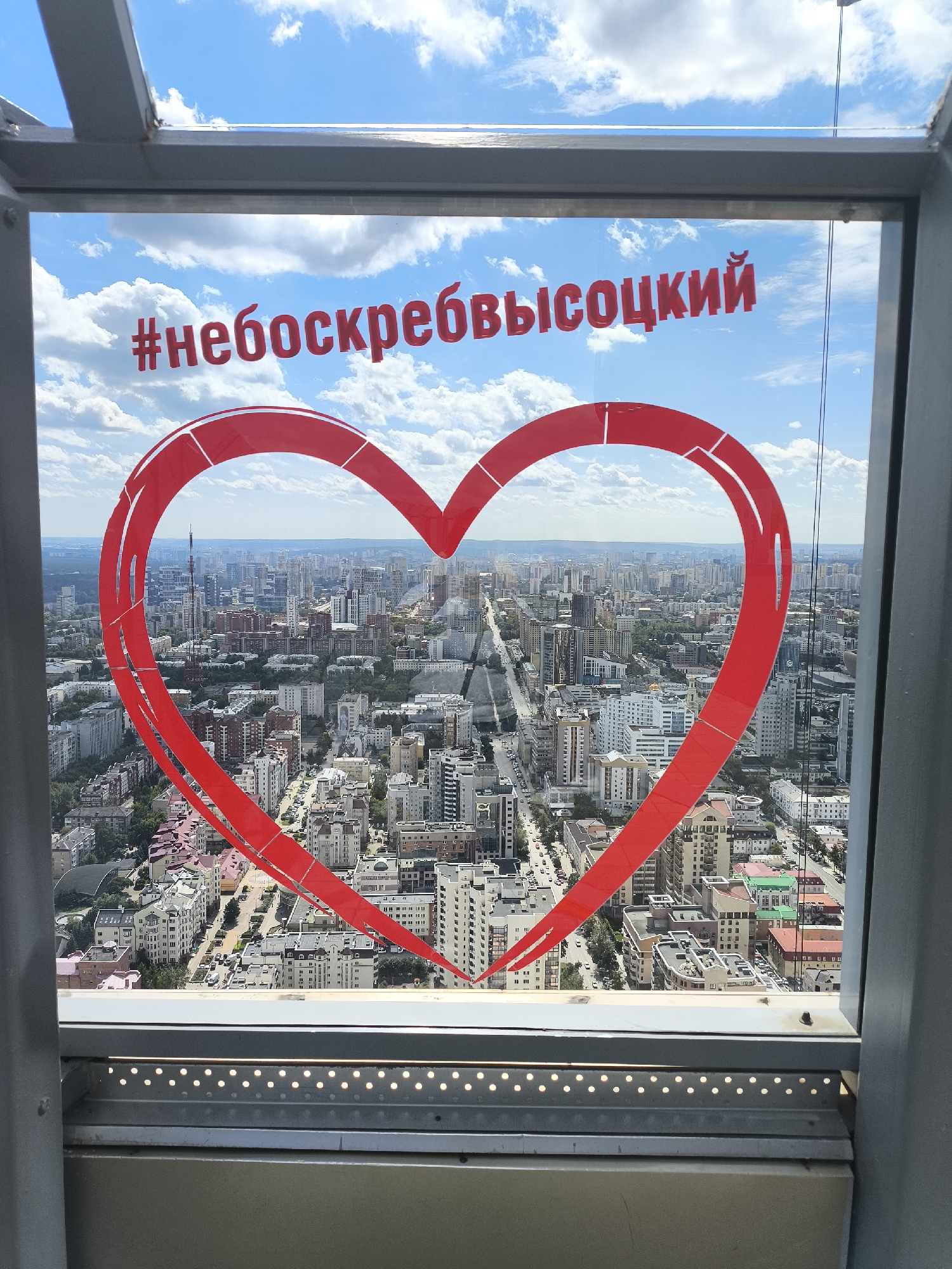The image is a photograph taken from a high vantage point, looking out through a window over a city. A large, red heart graphic is superimposed on the window, partially obscuring the cityscape below. Above the heart, the Russian hashtag "#небоскребывысоцкий" is written in red, block letters. The hashtag translates to "#skyscrapersvysotsky," likely referring to the Vysotsky business center in Yekaterinburg, Russia, known for its observation deck. The cityscape below is dense with buildings of various sizes and architectural styles, suggesting an urban environment. The sky is blue with scattered white clouds. The foreground shows the metal frame of the window and some lighting elements. No people are visible in the image.