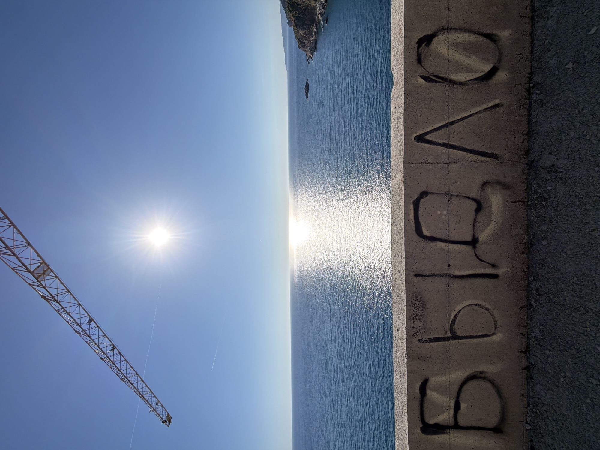 The image shows a coastal view from Perazića Do, Montenegro, with the Adriatic Sea dominating the scene. A concrete barrier runs along the right side of the frame, bearing graffiti that spells "Bravo." The sun is positioned high in the sky, creating a bright glare on the water's surface. A construction crane is visible in the lower left corner of the frame. In the distance, land is visible on the horizon, suggesting a coastline or islands.