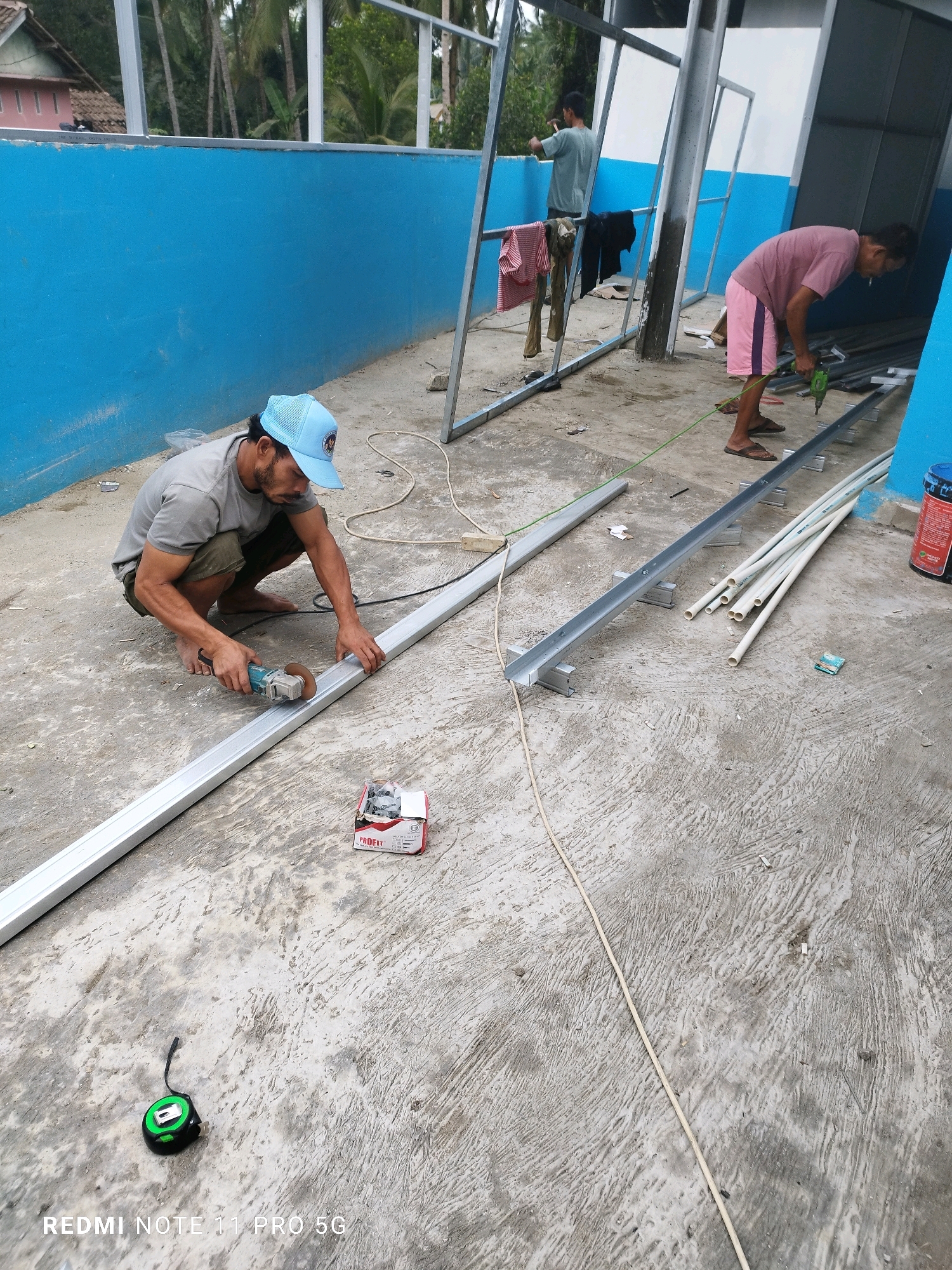In Sindangkarya, Indonesia, two men are working on a construction project. The man in the foreground, wearing a blue cap and a grey t-shirt, is crouched down and using an angle grinder to cut a long metal beam. He is barefoot and wearing shorts. In the background, another man in a pink t-shirt and pink shorts is using a green power drill to work on a metal frame. He is wearing sandals. The area appears to be outdoors, with a blue wall on the left and a roof structure overhead. Various construction materials, including pipes and metal bars, are scattered on the concrete ground. The overall scene depicts an active construction site.