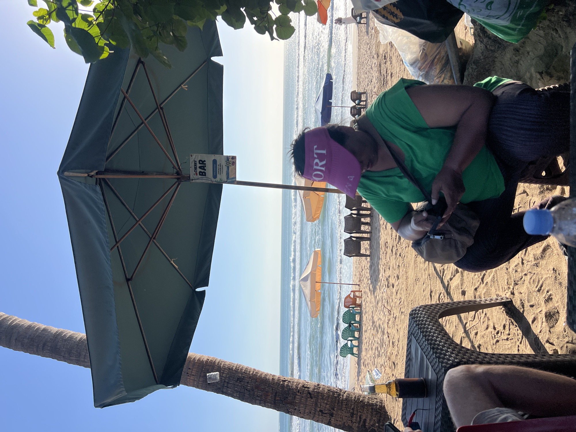 A woman wearing a pink visor and a green t-shirt is sitting on a stool on a beach in Kuta, Indonesia. She is looking down at her phone, which she is holding in her hands. The beach is sandy, and there are palm trees and umbrellas in the background. The ocean is calm, and there are waves crashing on the shore. There are also several chairs and tables on the beach, as well as a sign that says "CHERRY BAR." The sun is shining, and the sky is clear.