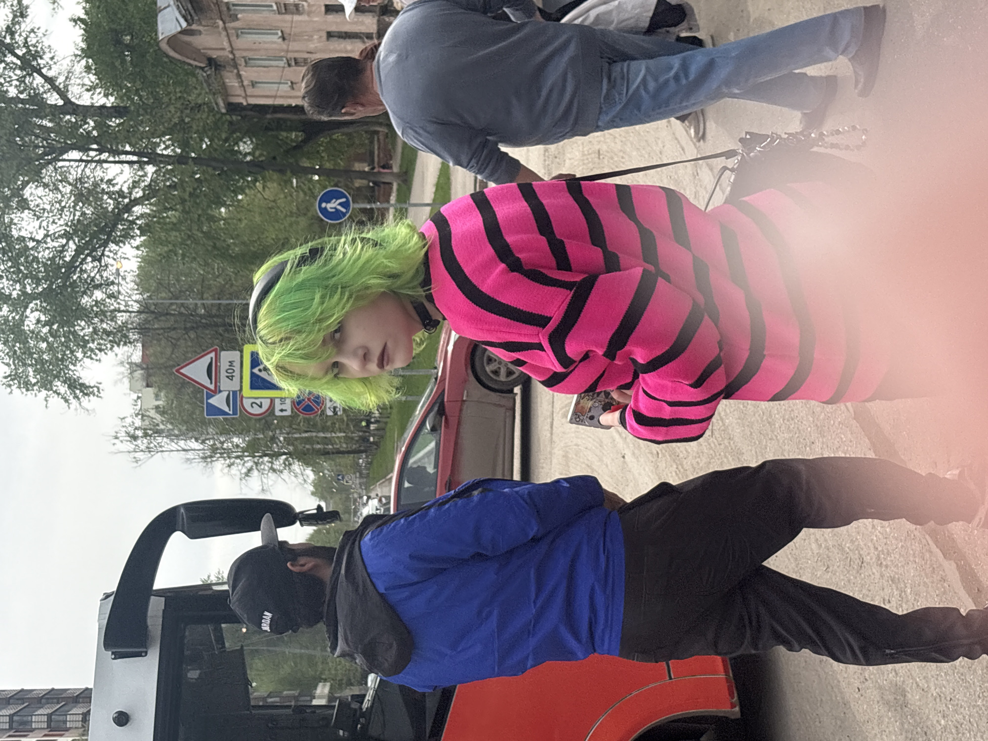 The image captures an outdoor scene in Perm, Russia, identified by signage. A person with bright green hair, wearing a black choker and a pink and black striped sweater, is visible in the foreground. A man in a blue jacket and black pants stands nearby, partially obscured by what appears to be a red bus. Another man in a grey shirt and jeans is walking a dog on a leash in the background. Traffic signs, including one indicating a speed limit and a pedestrian crossing sign, are visible. Buildings and trees line the background, suggesting an urban environment. The photo is taken from a low angle.