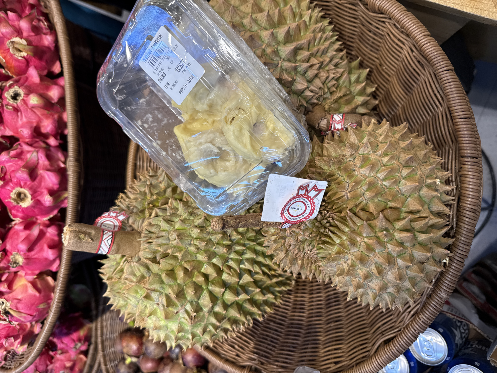 The image depicts a basket in what appears to be a retail setting, possibly in Kutuh, Indonesia given the reference to "Pepito Kutuh" on a packaged product. The basket contains several durian fruits, both whole and pre-packaged. The durians have a thorny exterior and some have price tags from “DURIAN MONTHONG BALI”. A clear plastic container holds durian fruit pieces. Adjacent to the basket are other fruits, including dragon fruit.
