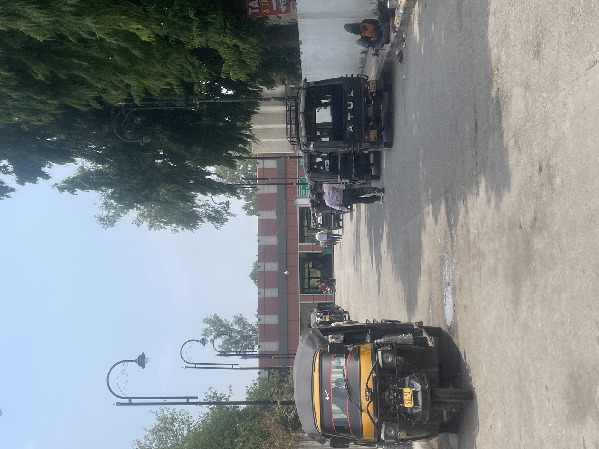 The image depicts a street scene in Pathankot, India, likely near a transportation hub such as a bus or train station, indicated by the presence of multiple auto-rickshaws and a building with what appears to be a waiting area or entrance.
In the foreground, a yellow and black auto-rickshaw is parked, its front end prominent. Behind it, a line of black auto-rickshaws is visible, some with passengers or drivers. Further down the street, several people are walking, including a person in a light-colored shirt and dark pants walking away from the camera.
The environment is characterized by a paved road and a sidewalk. Along the right side of the road, a white wall runs the length of the scene, with a sign partially visible at the far right. A person is seated on the curb next to the wall, appearing to be looking at a mobile phone. Adjacent to this person is a small pile of belongings and a container.
Overhead, decorative streetlights with curved arms are spaced along the street. Tall, leafy trees line the right side of the street, casting shadows on the road. The building in the background features a red and grey striped facade with arched entrances. The sky is clear and bright, suggesting a sunny day. The overall impression is one of a busy, public urban space.