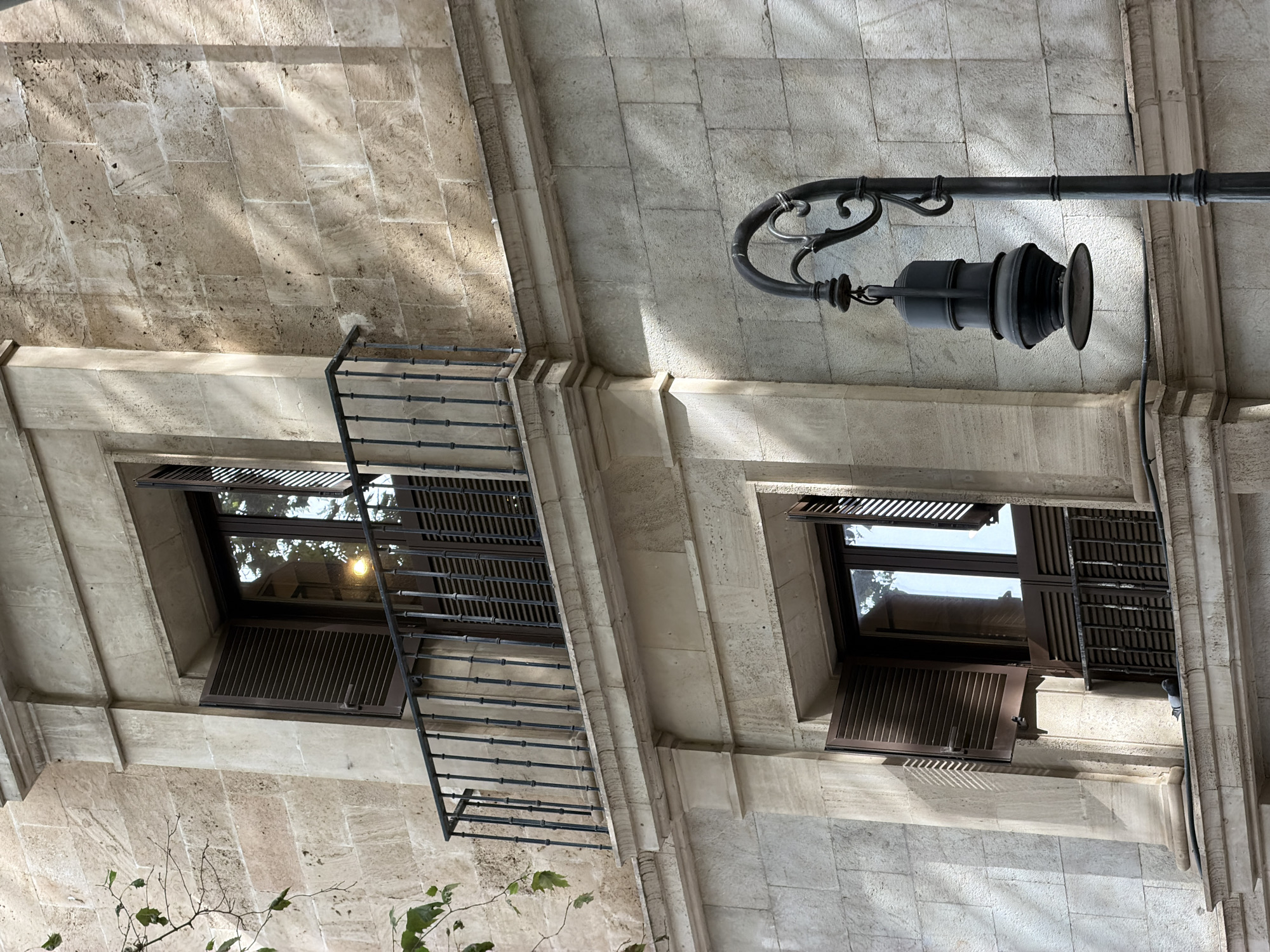 The image captures a detail of the exterior facade of a stone building, likely an older structure in Palma de Mallorca, Spain, given the architectural style. Two windows with dark wooden shutters are visible. The upper window features a small balcony with a black wrought-iron railing. Both windows have their shutters partially open, revealing a dark interior. A decorative black lamppost with an ornate curve and a cylindrical lantern hangs in the foreground to the right. Sunlight casts distinct shadows across the stone blocks of the building. A small branch with a few green leaves is visible on the left side of the image, extending from the upper left corner. There are no people present in the scene.