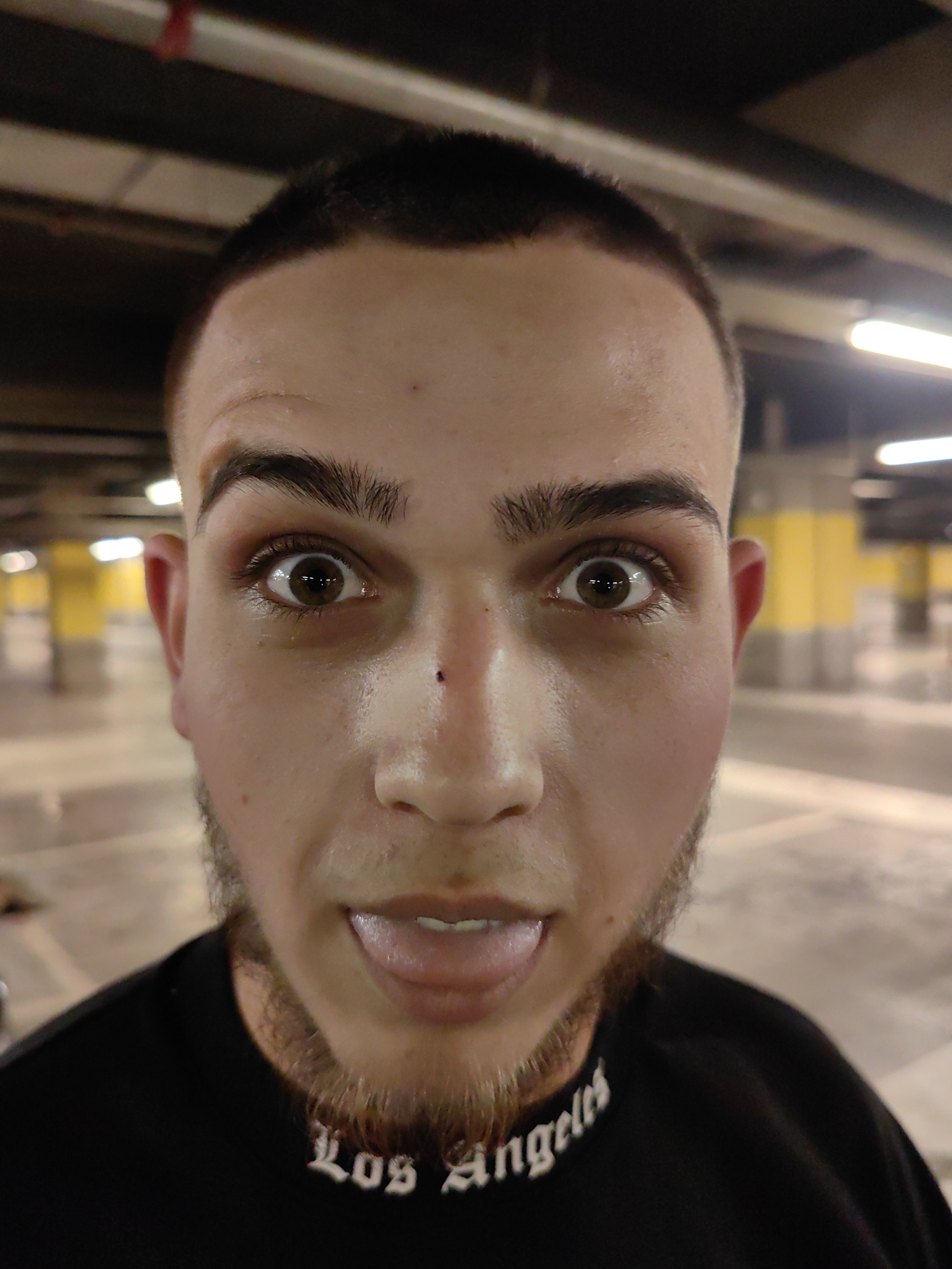 A medium-complex image depicts a young male individual, photographed from a low-angle perspective. His face dominates the foreground, showcasing his brown eyes, short, dark hair, and stubble beard. His tongue is slightly sticking out. He wears a black T-shirt with white "Los Angeles" script around the collar. The background reveals an indoor parking garage or similar enclosed space, with visible support columns and overhead lighting. The image suggests an intimate, close-up portrait taken in an urban, utilitarian setting. There is no specific indication that this is in Sofia, Bulgaria.