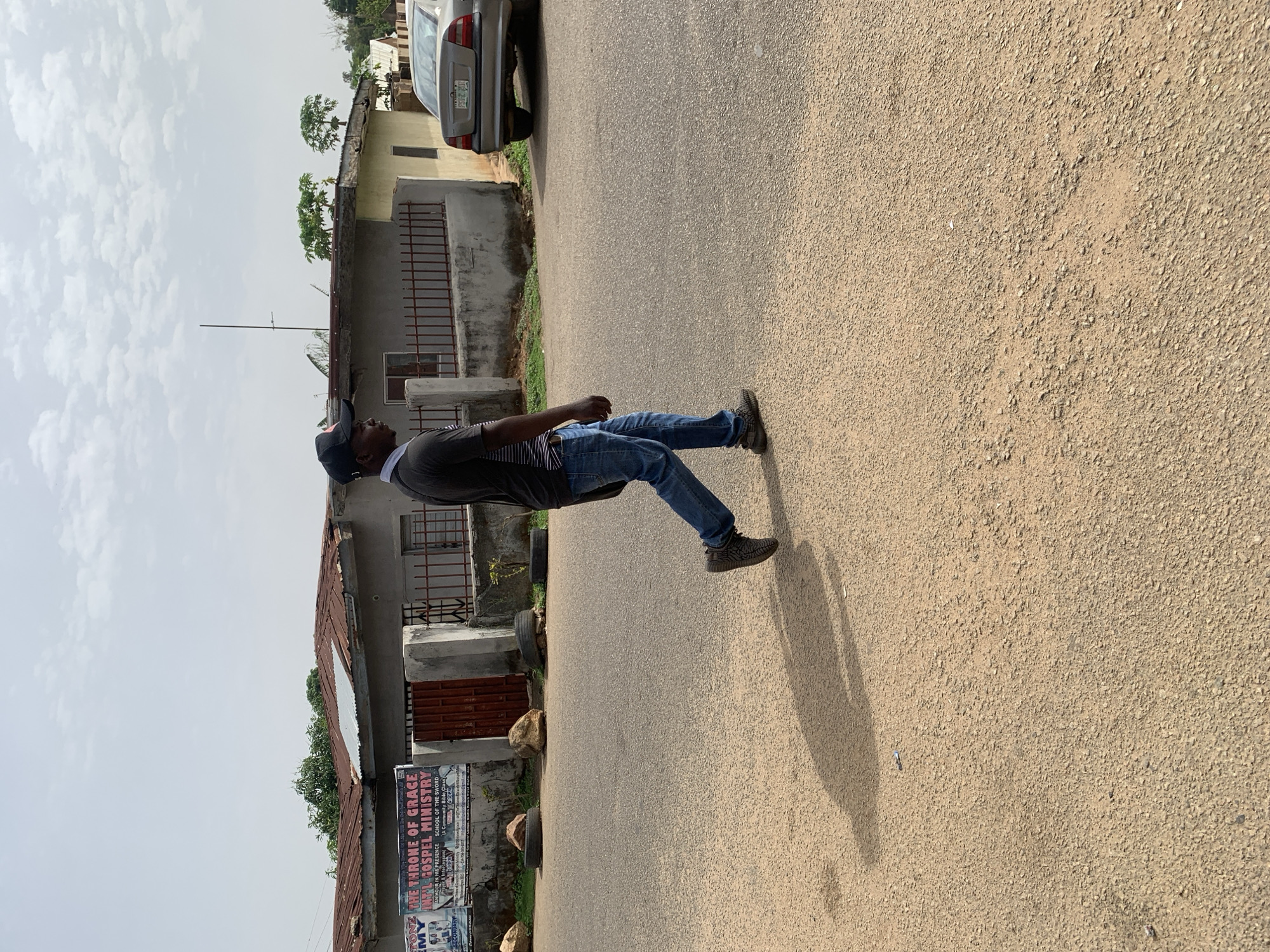 The image captures a man in casual attire (jeans, t-shirt, and cap) walking along a paved road in what appears to be a residential area. The location is Jos, Nigeria, indicated by the presence of a sign displaying "THE THRONE OF GRACE INT'L GOSPEL MINISTRY" on the side of a building. The environment includes buildings with corrugated iron roofs and fences, as well as a parked car. The road is visible with the man casting a shadow.